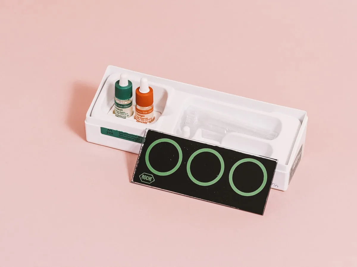 Una caja blanca con dos botellas pequeñas y una tarjeta negra con tres círculos verdes sobre una superficie rosa