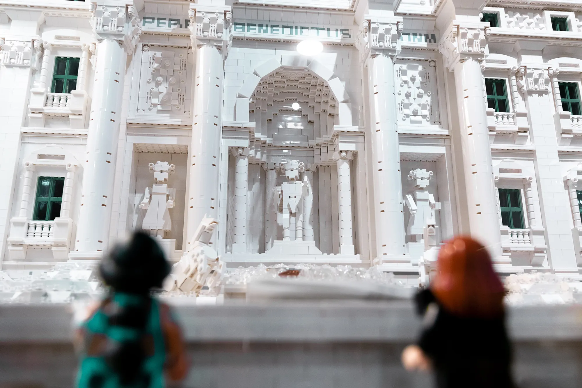 A LEGO model of a classical building with columns and statues, with two people observing it in the foreground
