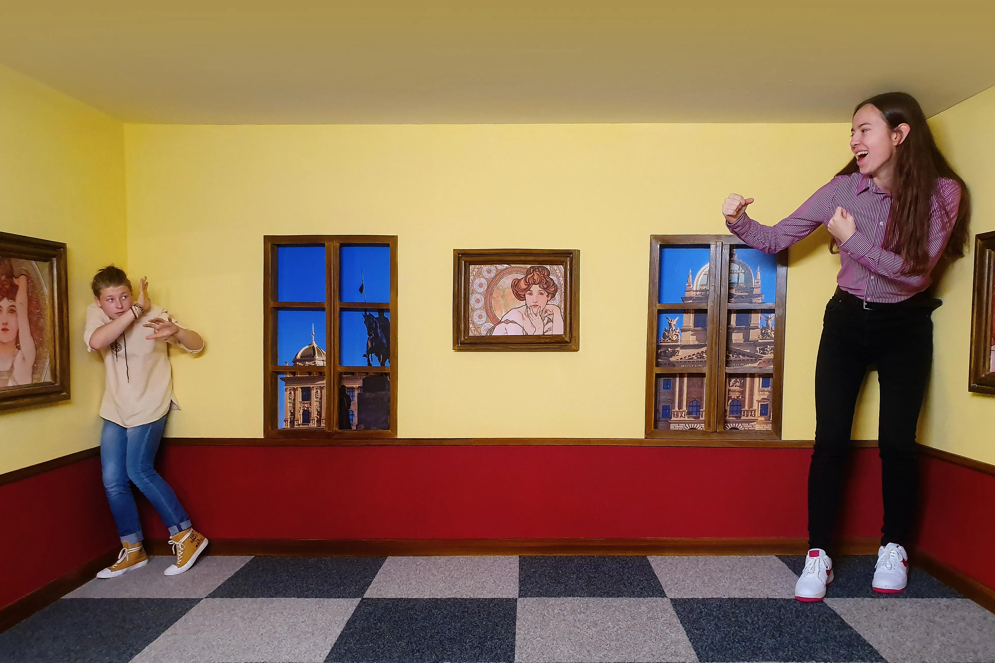 Two people posing in a room with yellow walls and a checkered floor, appearing distorted by an optical illusion through framed windows showing cityscapes.