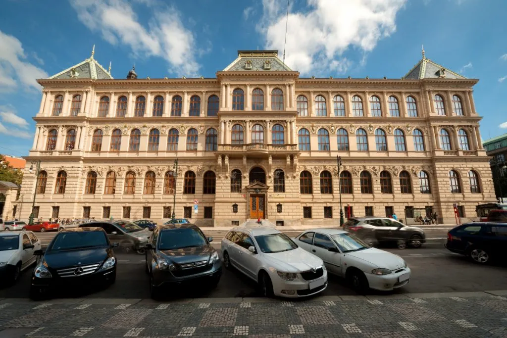Budynek neorenesansowy z łukowymi oknami i centralnym wejściem, otoczony zaparkowanymi samochodami na brukowanej ulicy.