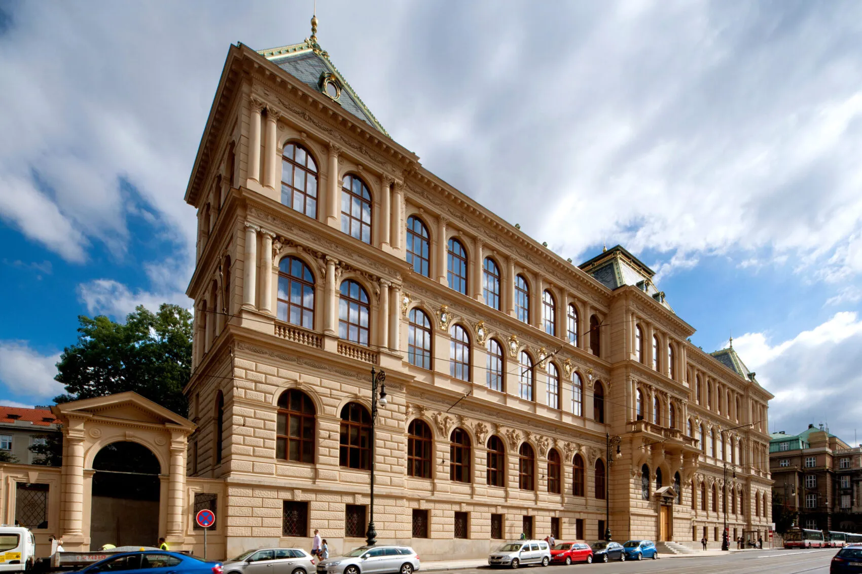 Budynek w stylu neorenesansowym z beżową kamienną fasadą, łukowymi oknami i centralną wieżą pod błękitnym niebem z chmurami