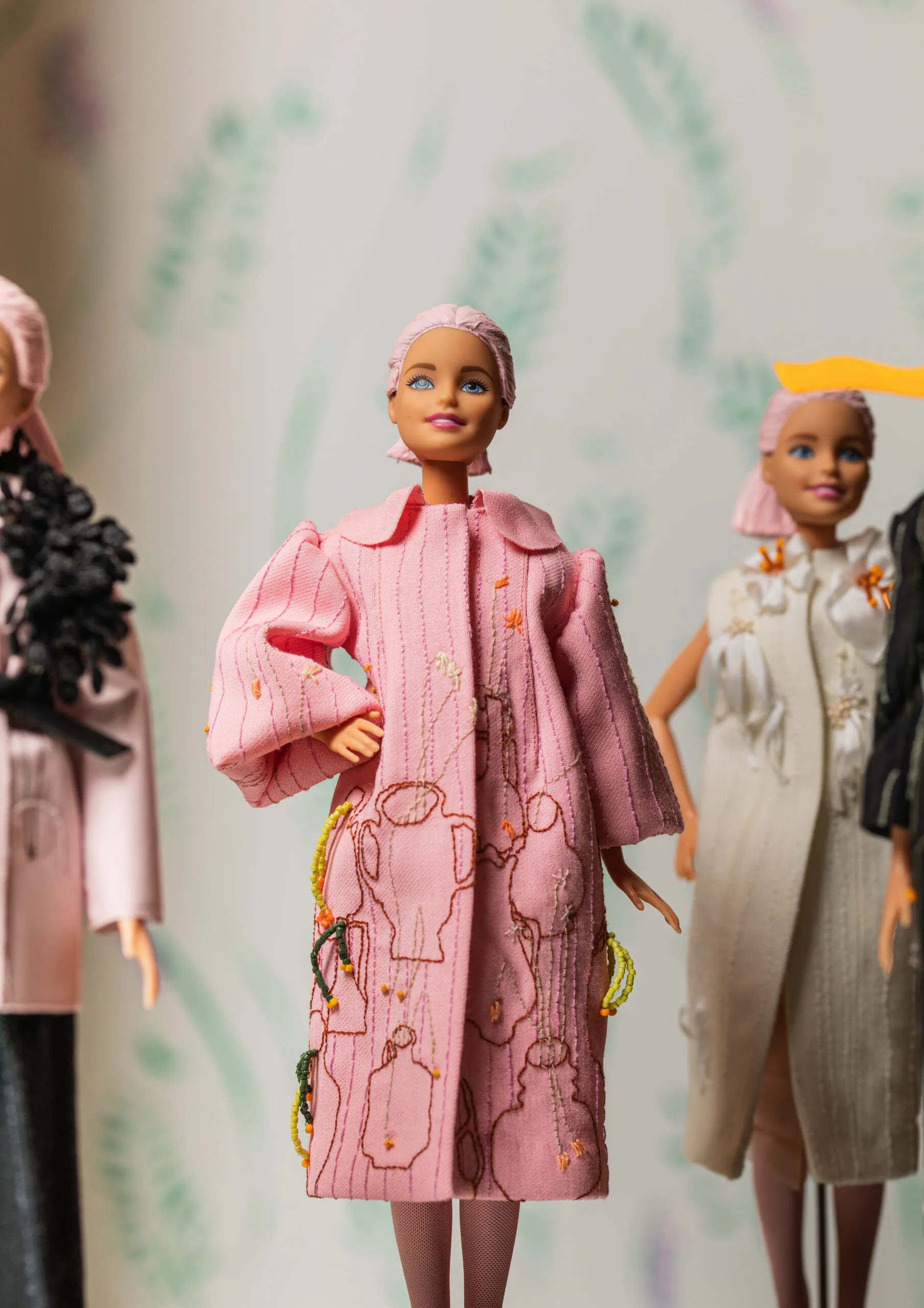 Lalki Barbie w zaprojektowanych na zamówienie strojach na wystawie, z centralną lalką w różowym, haftowanym płaszczu z ozdobnymi wzorami
