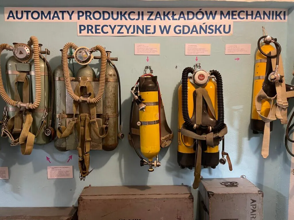 diving equipment including helmets, tanks, and hoses mounted on a wall with a sign in Polish