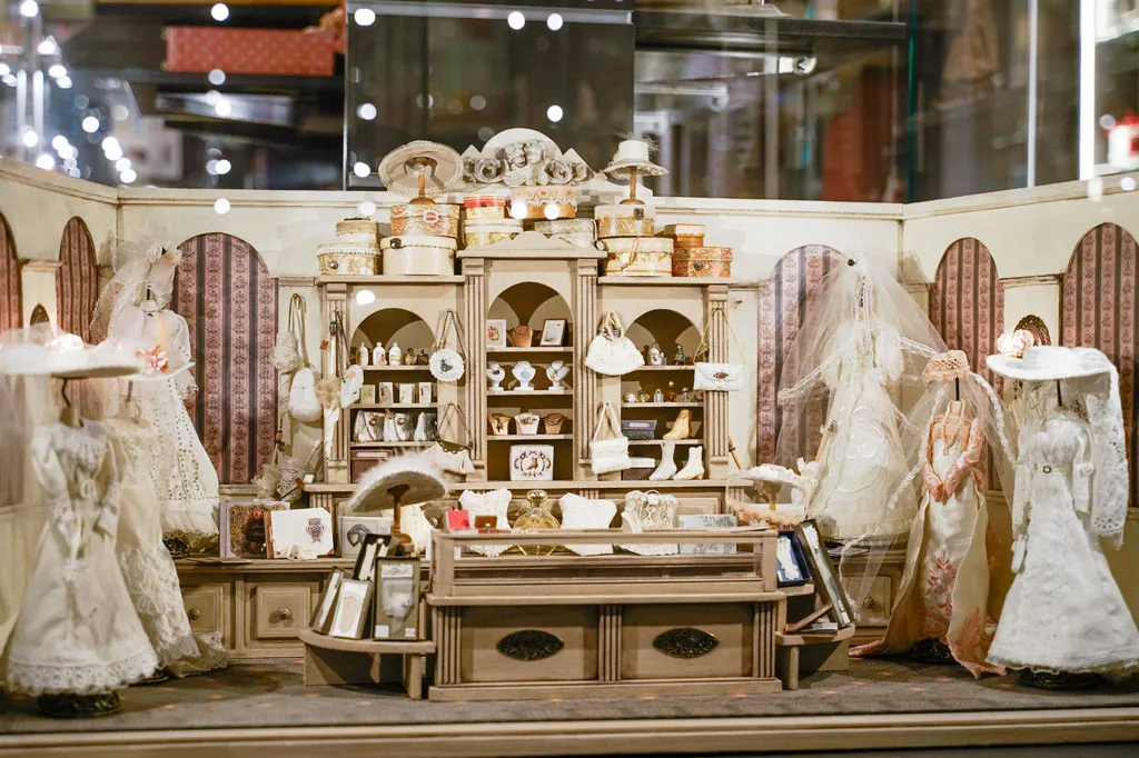 A museum display case featuring miniature dollhouses, toys, and mannequins in white dresses.