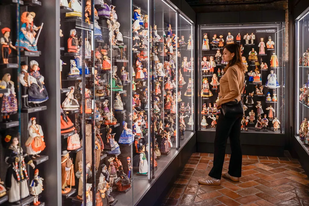 A woman stands in a museum gallery looking at glass cases filled with dolls and toys.