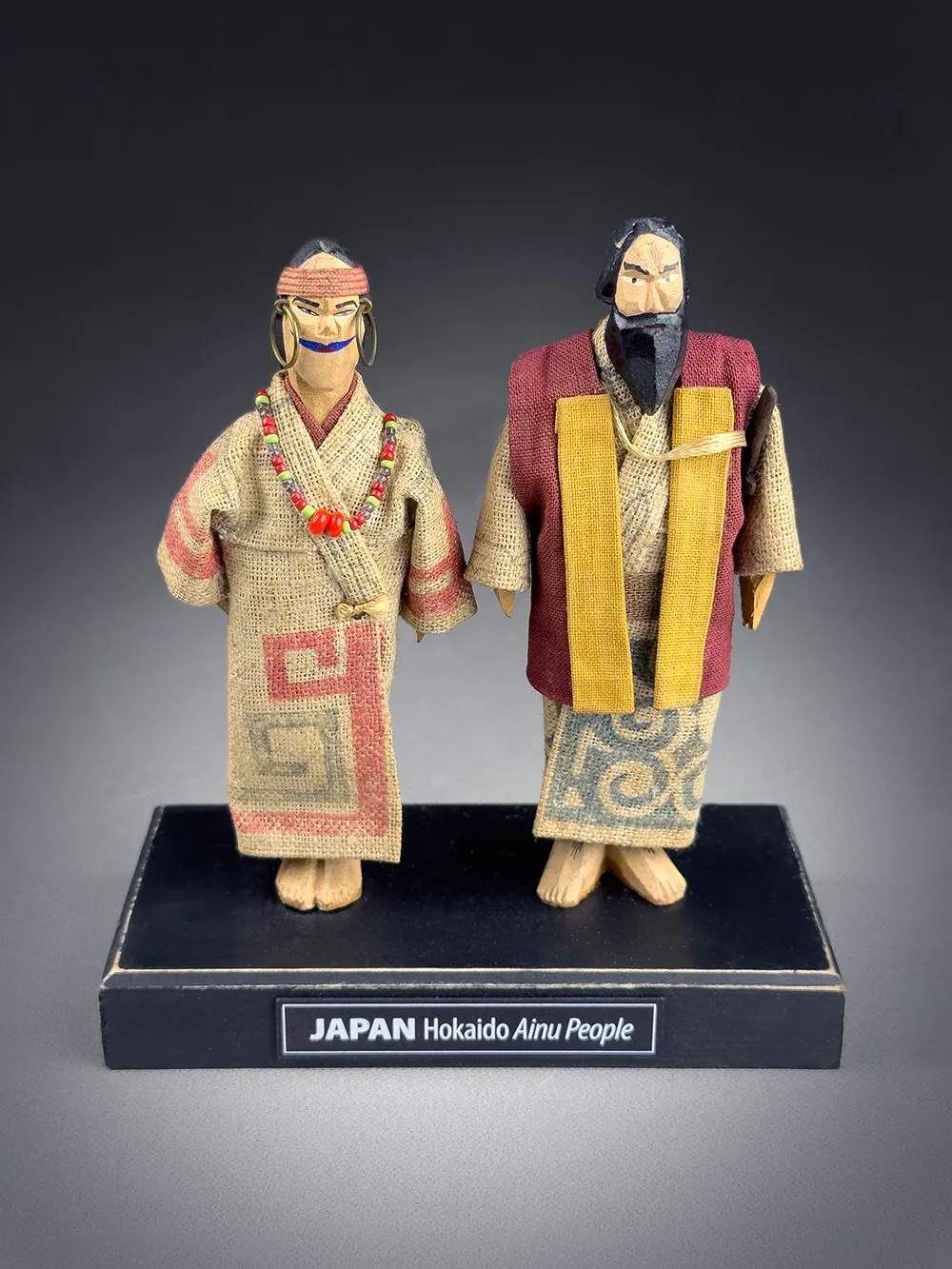 Two traditional dolls in Ainu attire displayed on a black base with a label reading 'JAPAN Hokkaido Ainu People'
