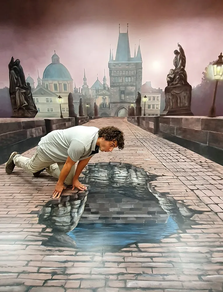 Mężczyzna czołgający się po brukowanej podłodze z namalowaną iluzją lśniącej kałuży, otoczony namalowanym miejskim krajobrazem praskiej architektury i rzeźb.