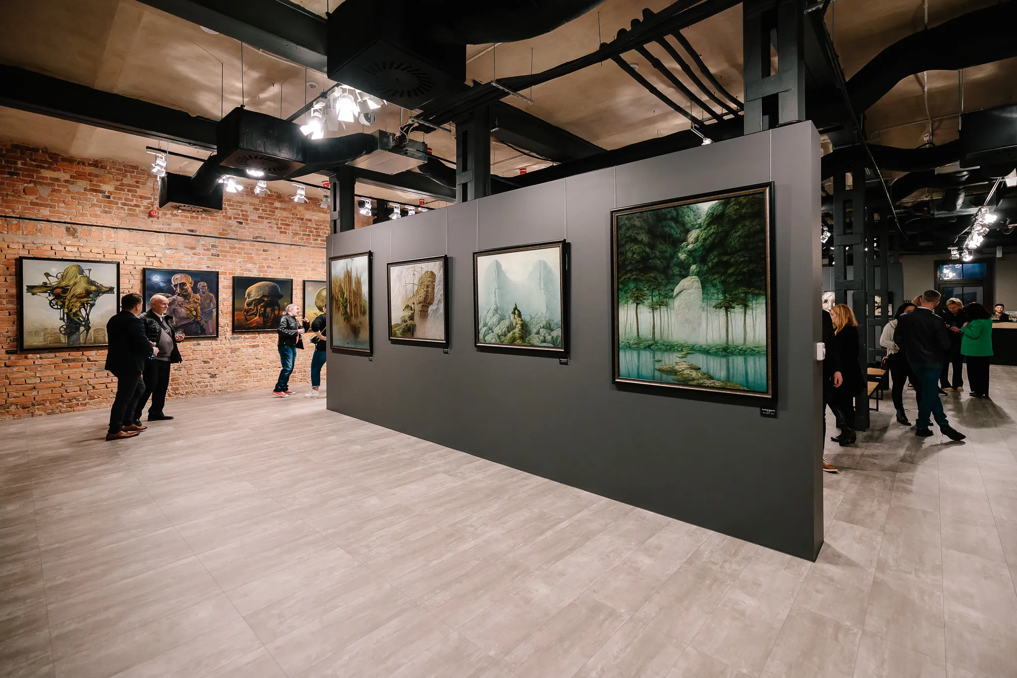 Galería de arte con pinturas enmarcadas en una pared gris, visitantes caminando, pared de ladrillo expuesto y techo industrial