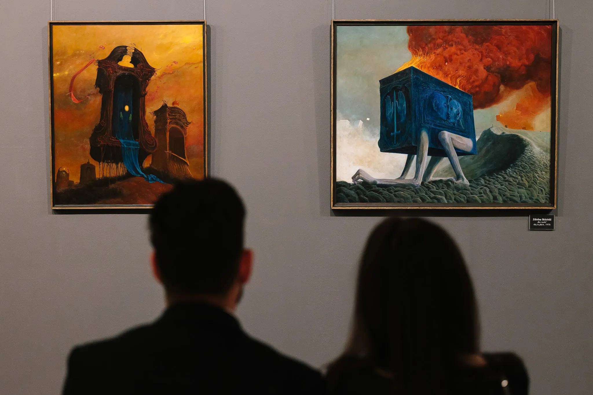 Dos personas observando pinturas surrealistas en una galería, una representando un castillo con una figura azul y la otra mostrando un cubo con patas contra un cielo ardiente.