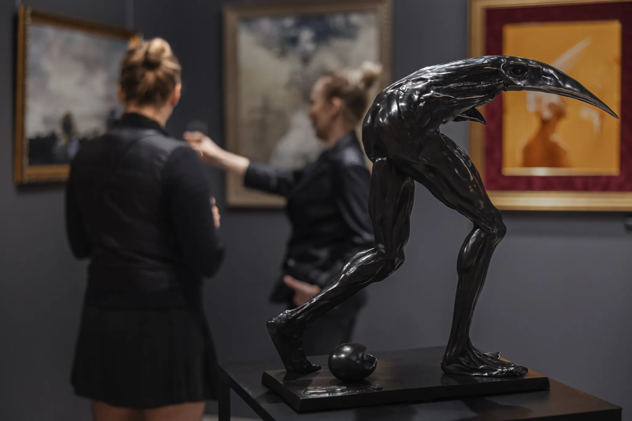 Una escultura oscura y brillante de una figura humanoide con extremidades alargadas y un pico parecido al de un ave, expuesta en una galería con visitantes observando y pinturas enmarcadas al fondo.