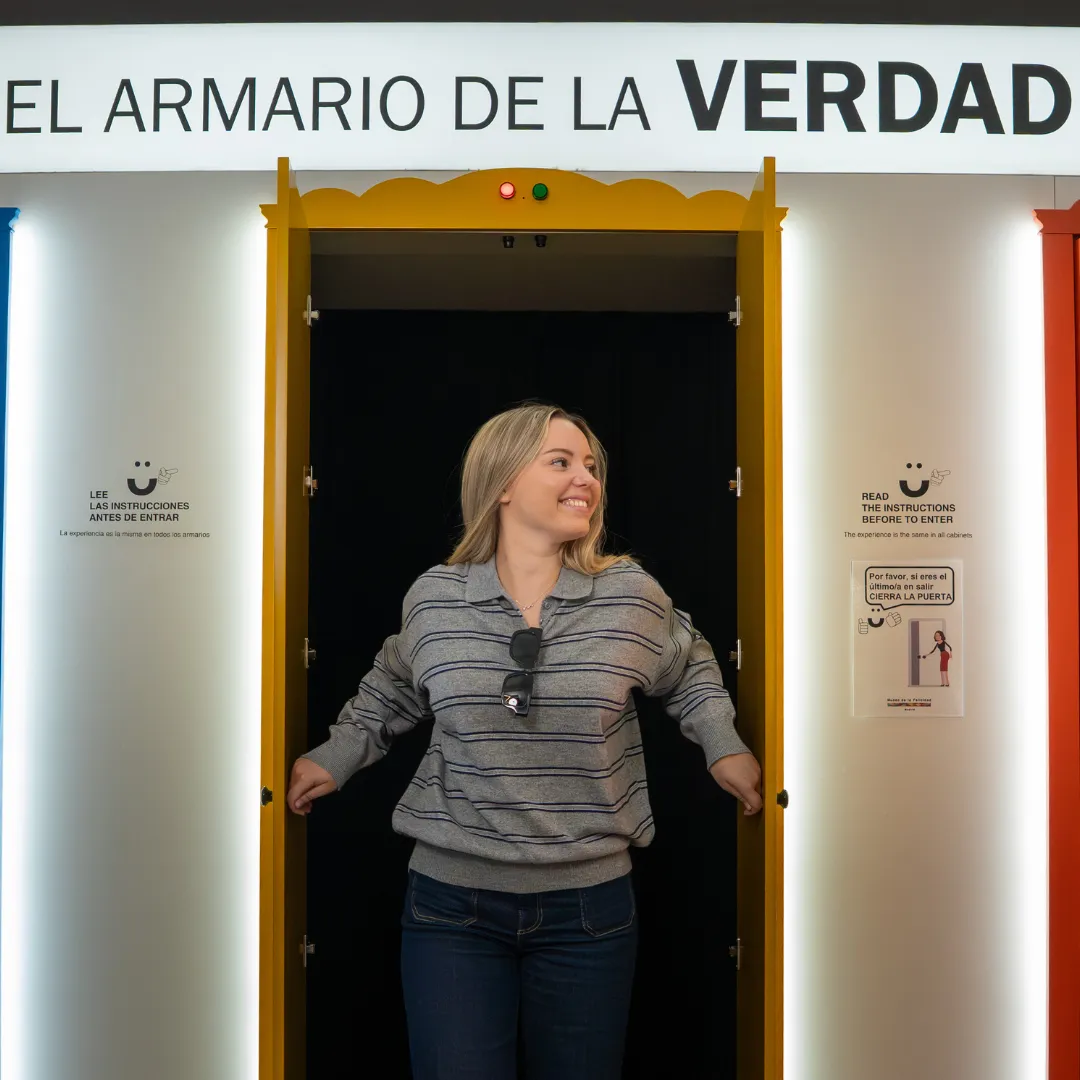 Een vrouw in een gestreepte blouse staat in een gele deuropening met een bord erboven waarop 'EL ARMARIO DE LA VERDAD' staat op een witte muur.