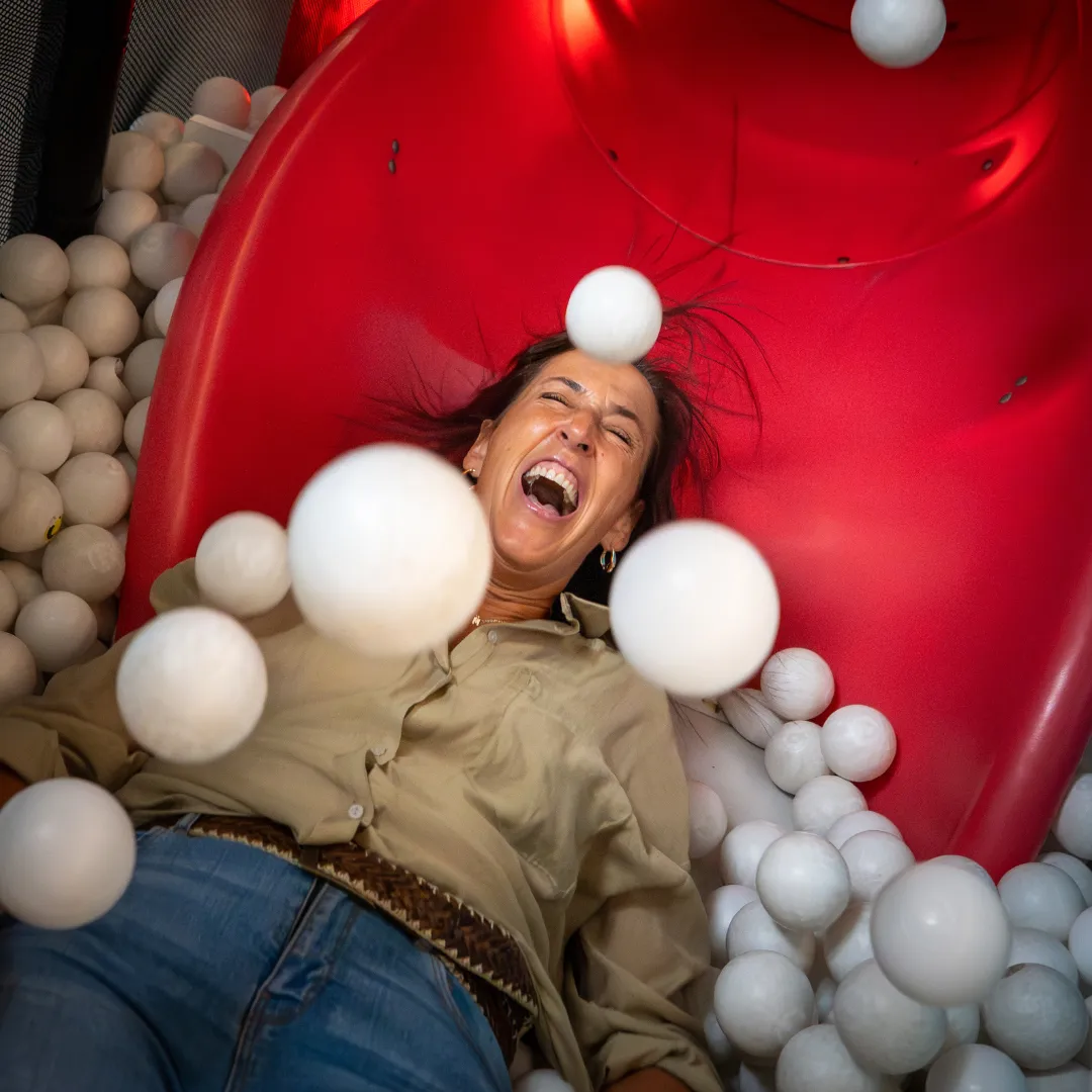 Een lachend persoon met gesloten ogen glijdt van een rode glijbaan omringd door witte ballen