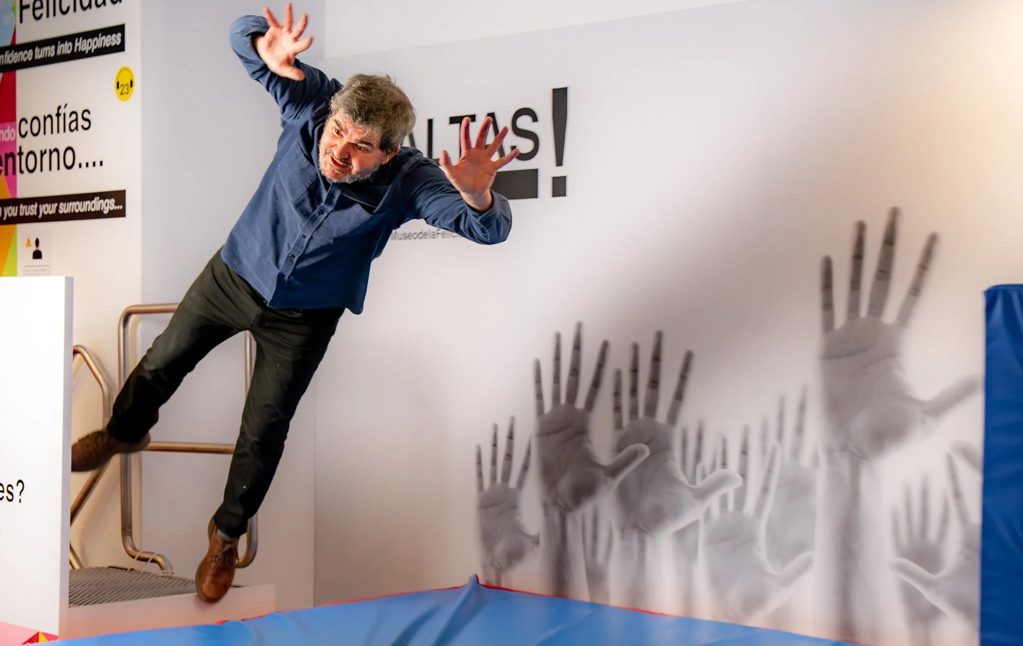 Een man die op een trampoline springt voor een muur met handafdrukken en het woord 'FELICIDAD!'