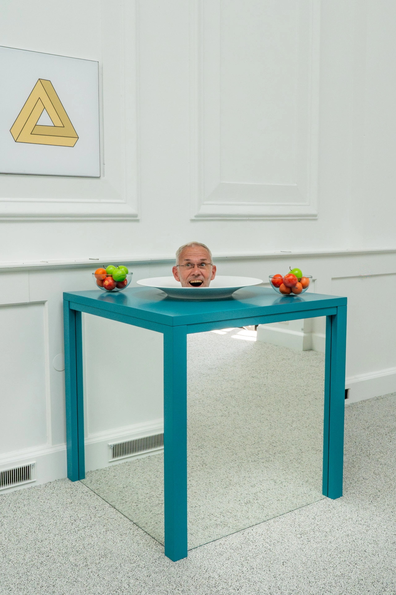 An optical illusion exhibit featuring a person's head appearing in a bowl on a blue table with mirrored base, fruit bowls, and an impossible triangle artwork on white paneled walls
