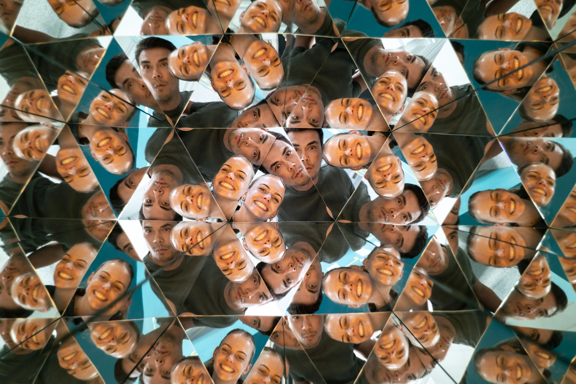 Kaleidoscopic mirror exhibit forming triangular reflections of multiple people's faces in symmetrical patterns.