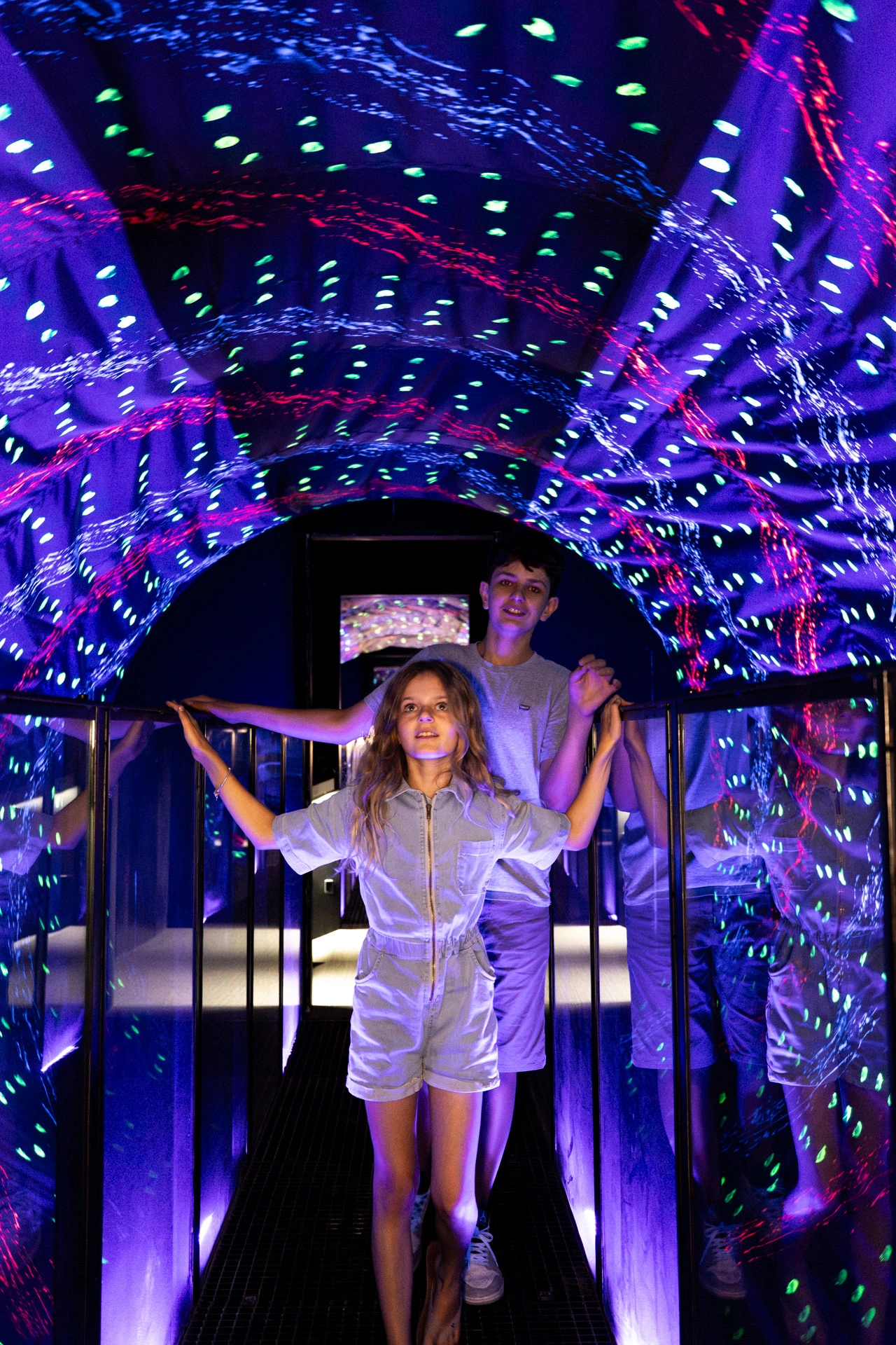 Two people experience a colorful LED-lit optical illusion tunnel with mirrored walls at the Museum of Illusions Amsterdam.