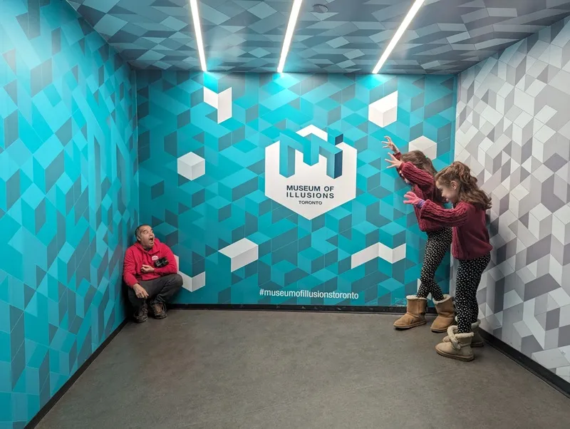 Children experiencing an optical illusion exhibit with geometric patterns on blue and gray walls and a Museum of Illusions logo