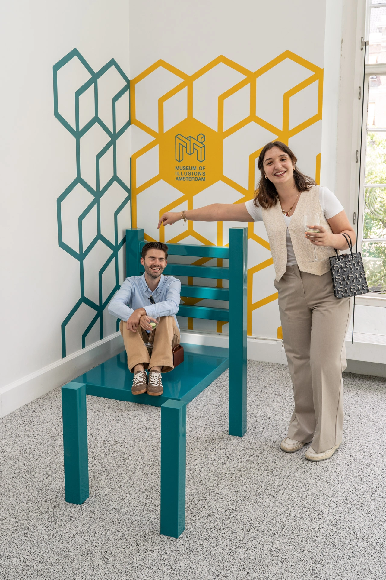 Two people interacting with a distorted perspective chair exhibit against a geometric patterned wall at Museum of Illusions Amsterdam