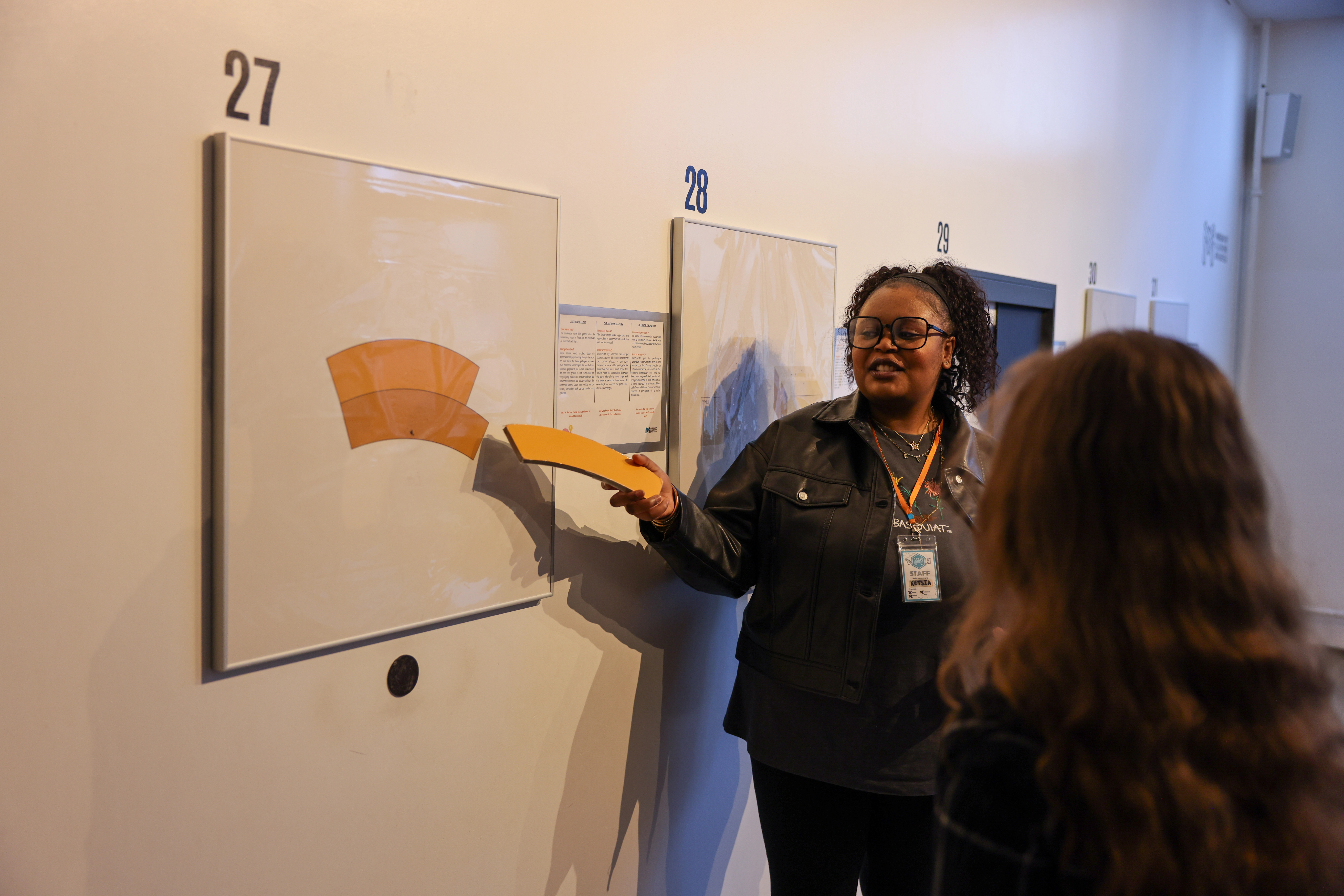 A woman demonstrates an optical illusion exhibit with a yellow curved shape at number 27, while another person observes in a museum gallery.