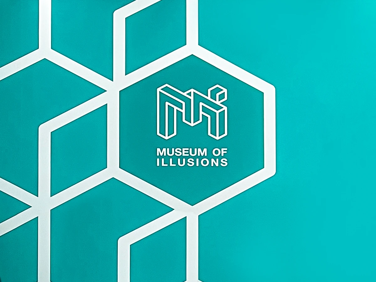 Białe logo Museum of Illusions ze stylizowanymi literami M i I w sześciokącie na turkusowym tle ze wzorem geometrycznym