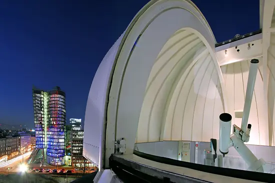 Biała, zakrzywiona konstrukcja architektoniczna Muzeum Iluzji Wiedeń nocą z oświetlonym drapaczem chmur w tle