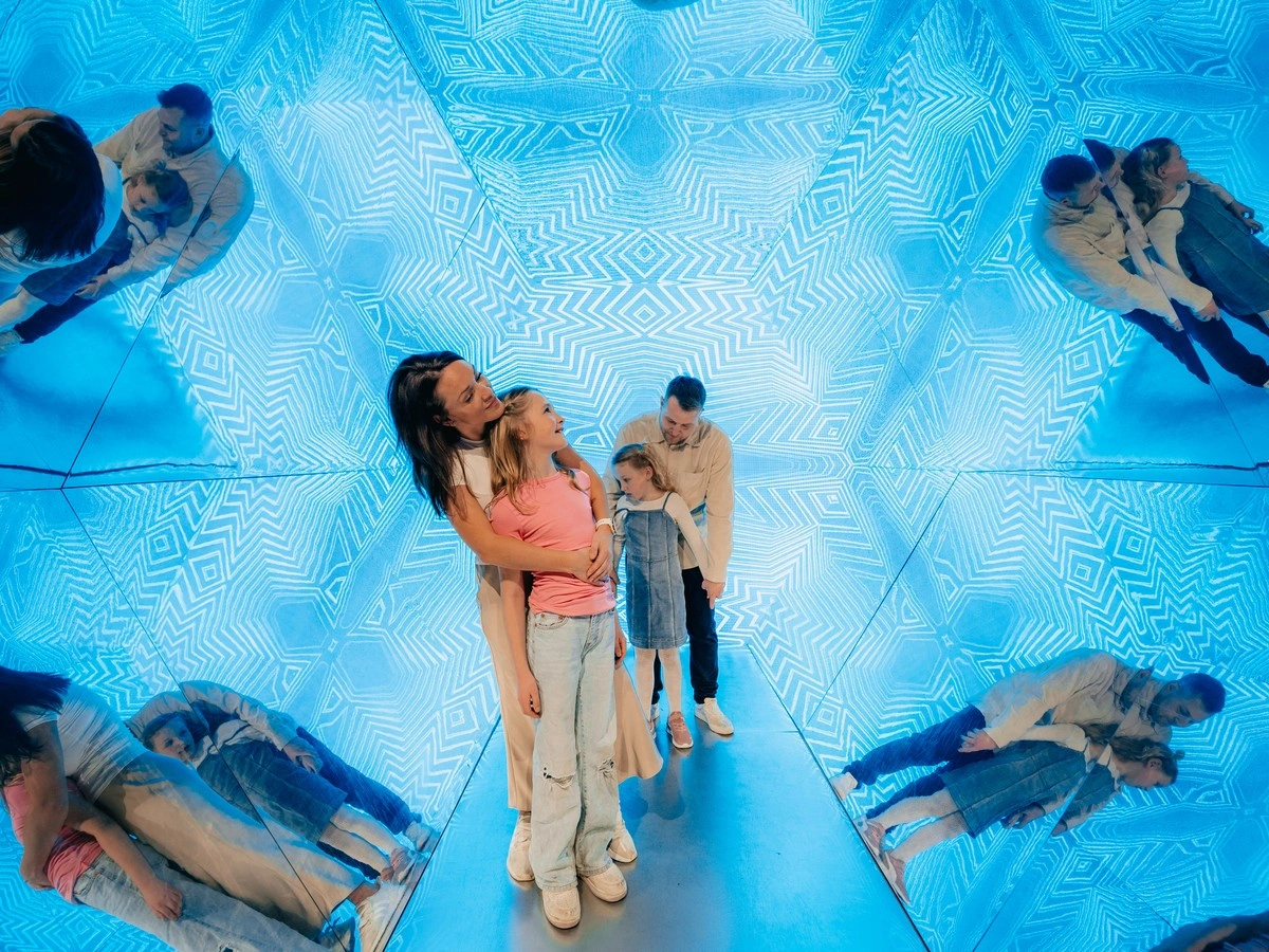 Een gezin van vier personen dat staat in een blauwe, patroonkamer met optische illusies die meerdere reflecties van hun figuren creëren.