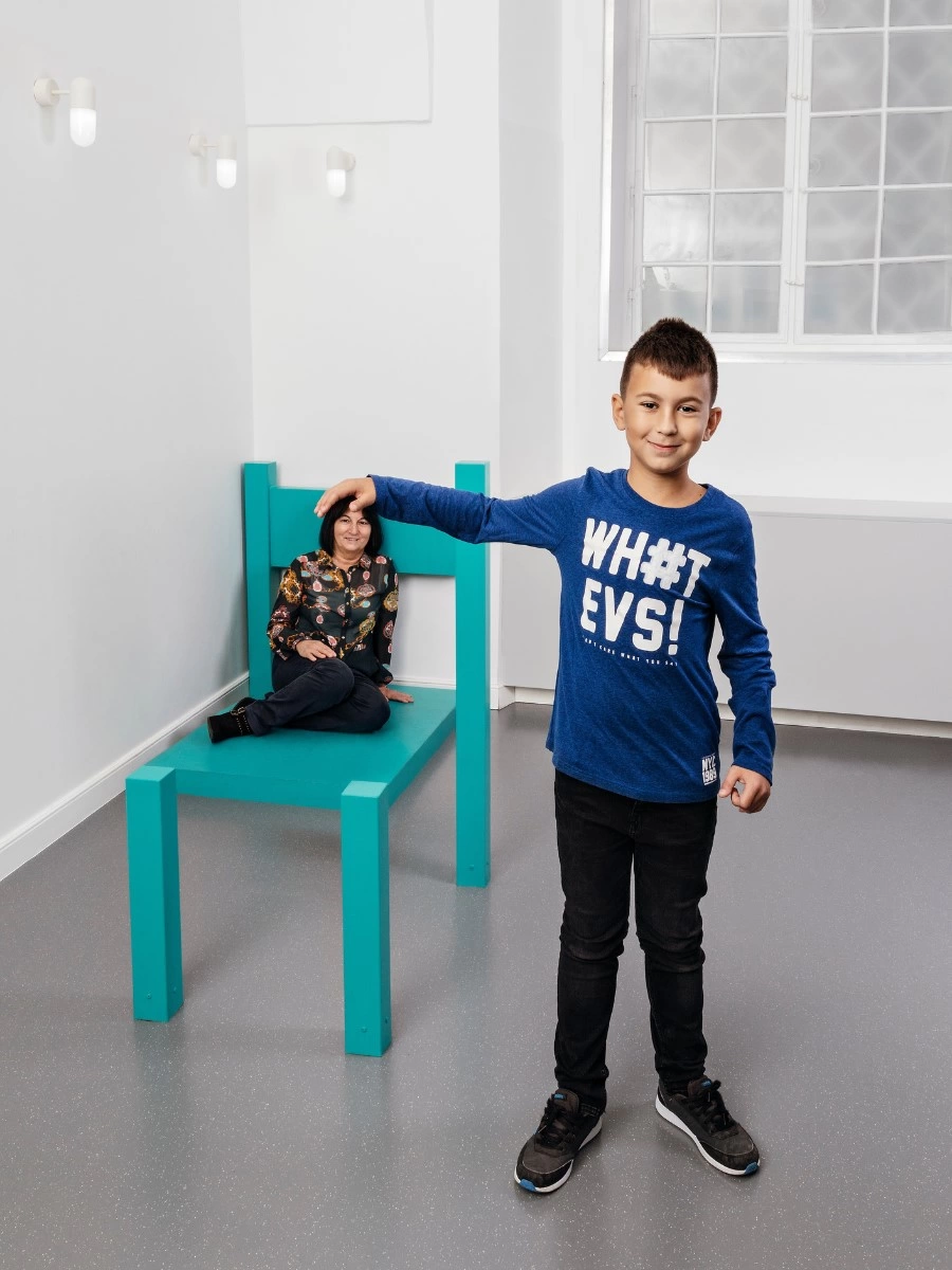 Een jongen in een blauw shirt staat naast een grote turkooizen stoel waar een vrouw in zit, wat een optische illusie van grootteverschil creëert in een witte kamer met een raam.