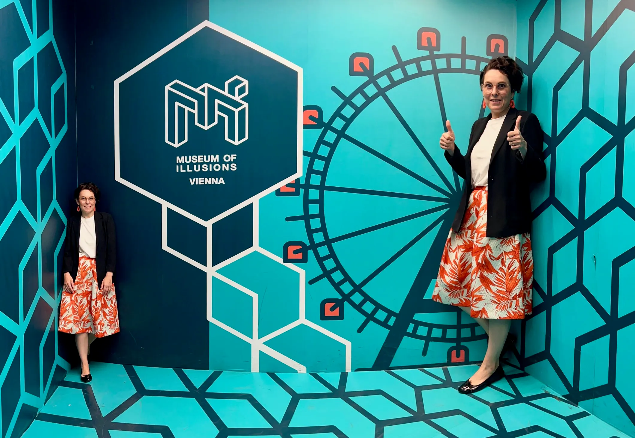 Twee vrouwen staan in een illusiekamer met een blauwe geometrische muur, een groot hexagonlogo van het Museum of Illusions Vienna en een rond optisch illusie-ontwerp.