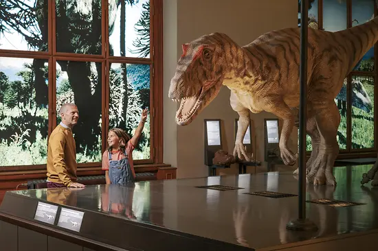 Duży model dinozaura za lustrzanym stołem z mężczyzną i dzieckiem obserwującymi go w galerii muzealnej