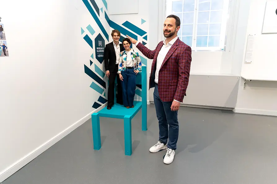 Drie mensen die staan in een illusietentoonstelling met een grote blauwe stoel en een geometrisch muurpatroon