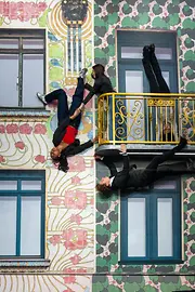 Twee mensen die van een balkon lijken te hangen aan een kleurrijk, patroon gevel