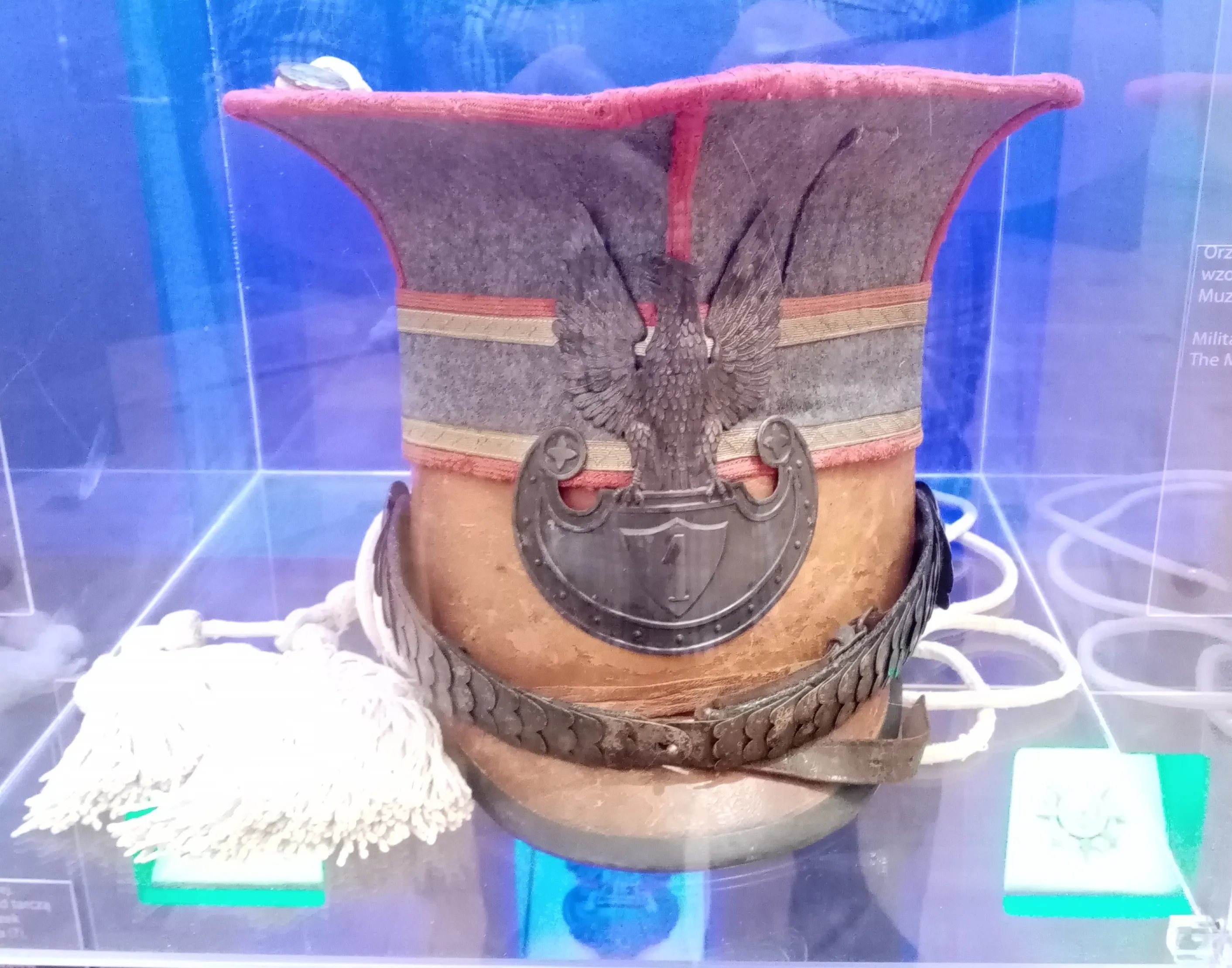 A historical military hat with a red top, gray fabric, and an eagle emblem, displayed in a glass case