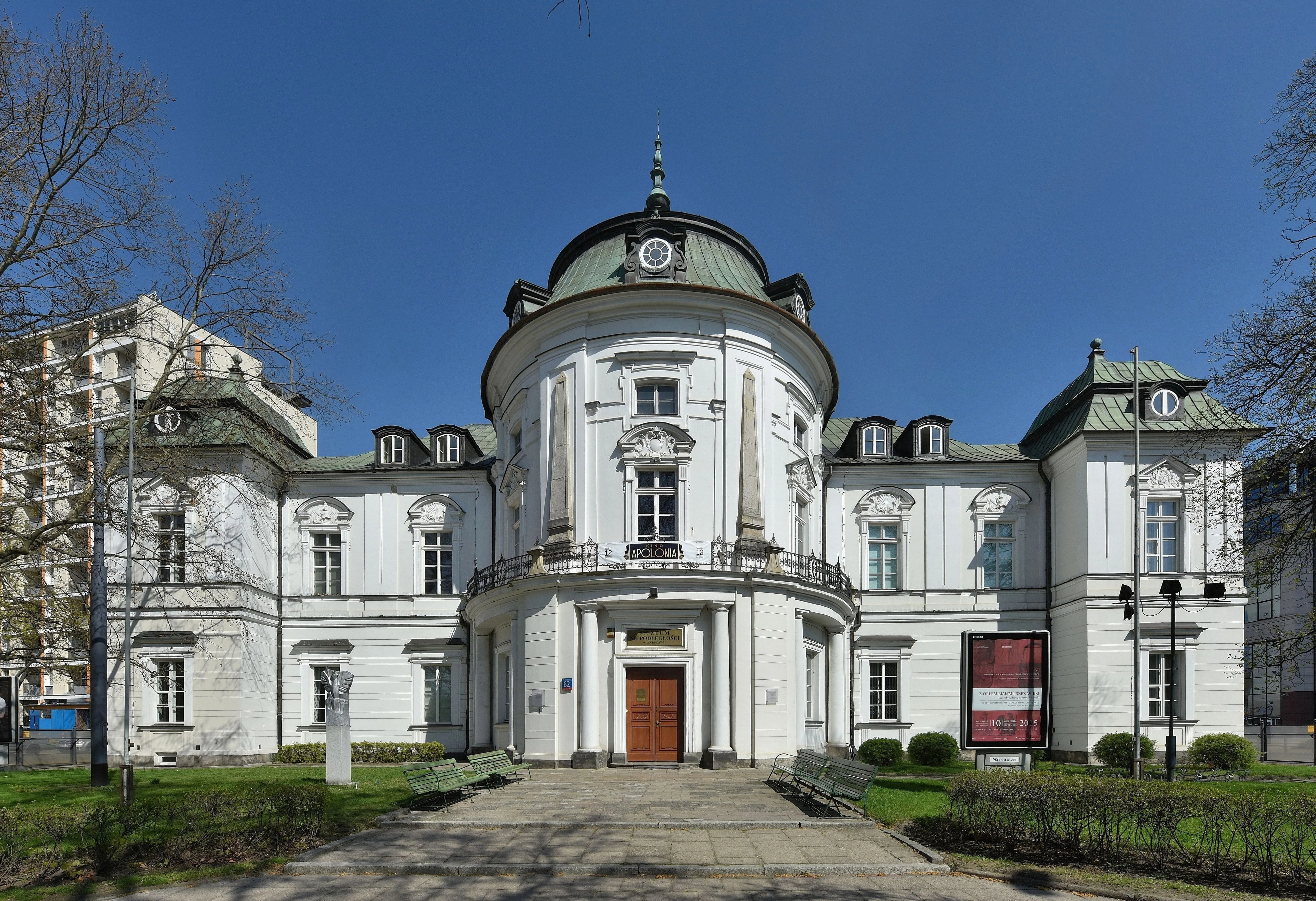 White classical building with a central dome and columns, green roof, and a red door entrance under a blue sky