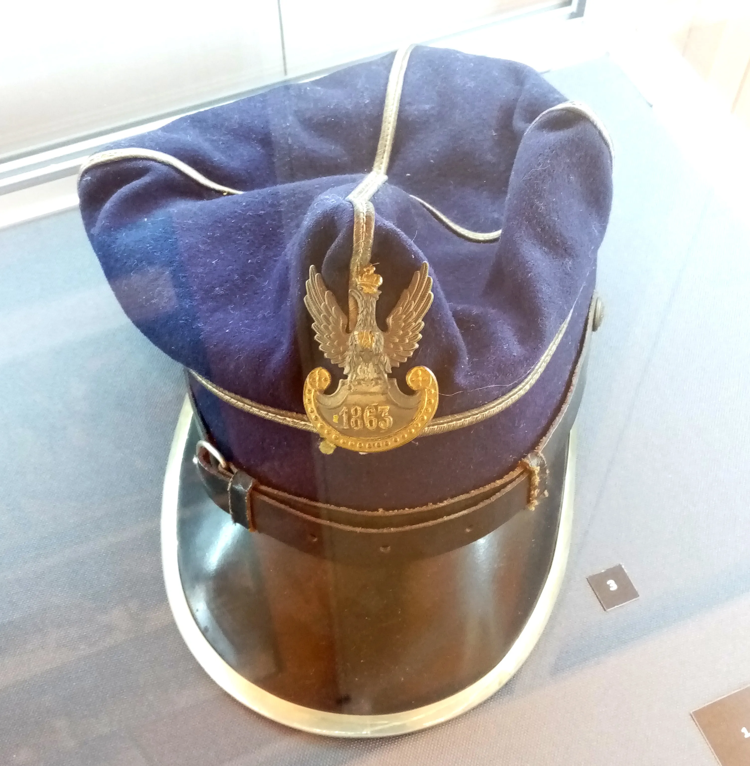 A blue military cap with a gold eagle emblem and the year 1863, displayed in a glass case