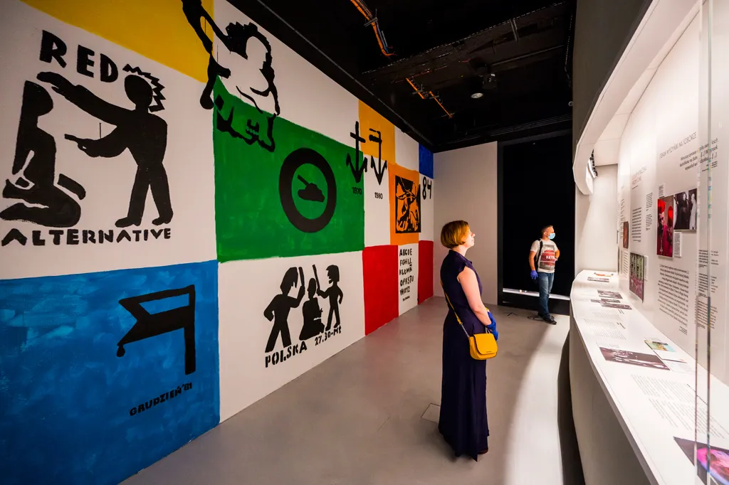 Een vrouw in een donkere jurk staat in een museumgalerij en kijkt naar een muur met een kleurrijke muurschildering met grafische silhouetten en tekst, waaronder 'RED ALTERNATIVE' en 'POLSKA BURZA'.