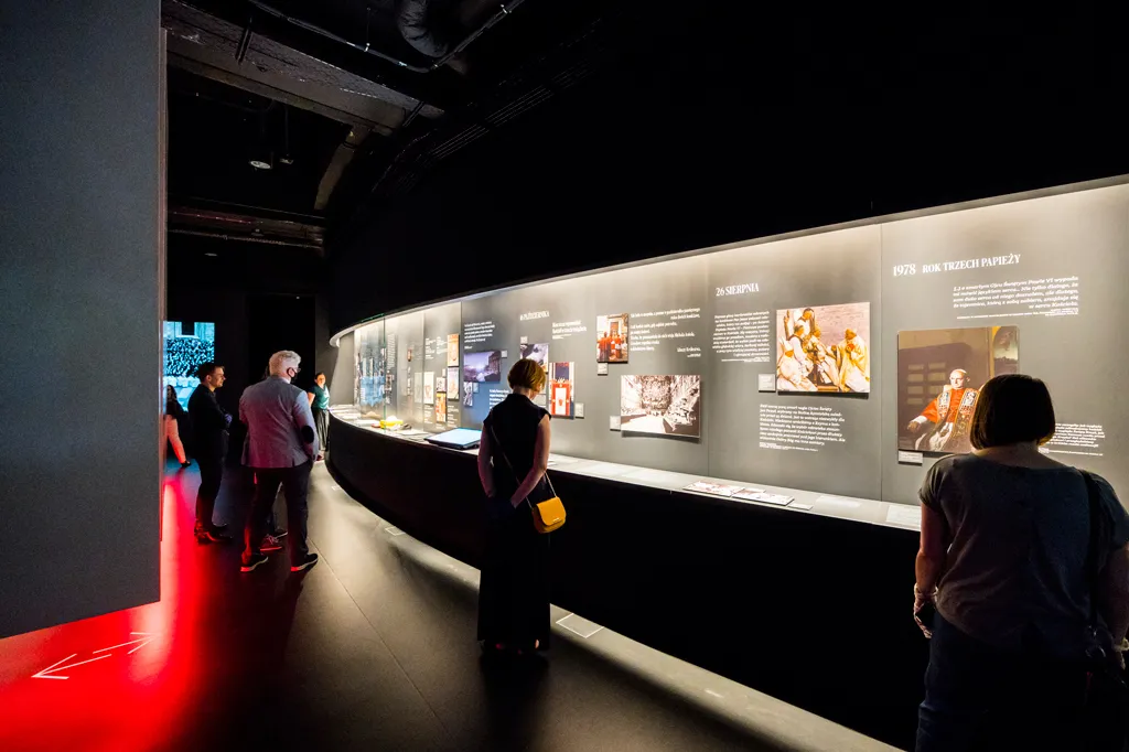 Tentoonstellingsruimte met bezoekers die een lange wand met multimediaprojecten en tekstpanelen bekijken onder gerichte verlichting.