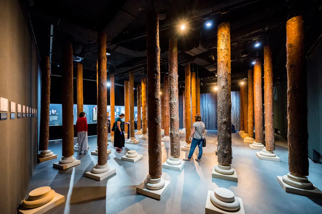 Interieur van een museum met hoge houten pilaren op witte voetstukken, bezoekers lopen door de ruimte onder spots.