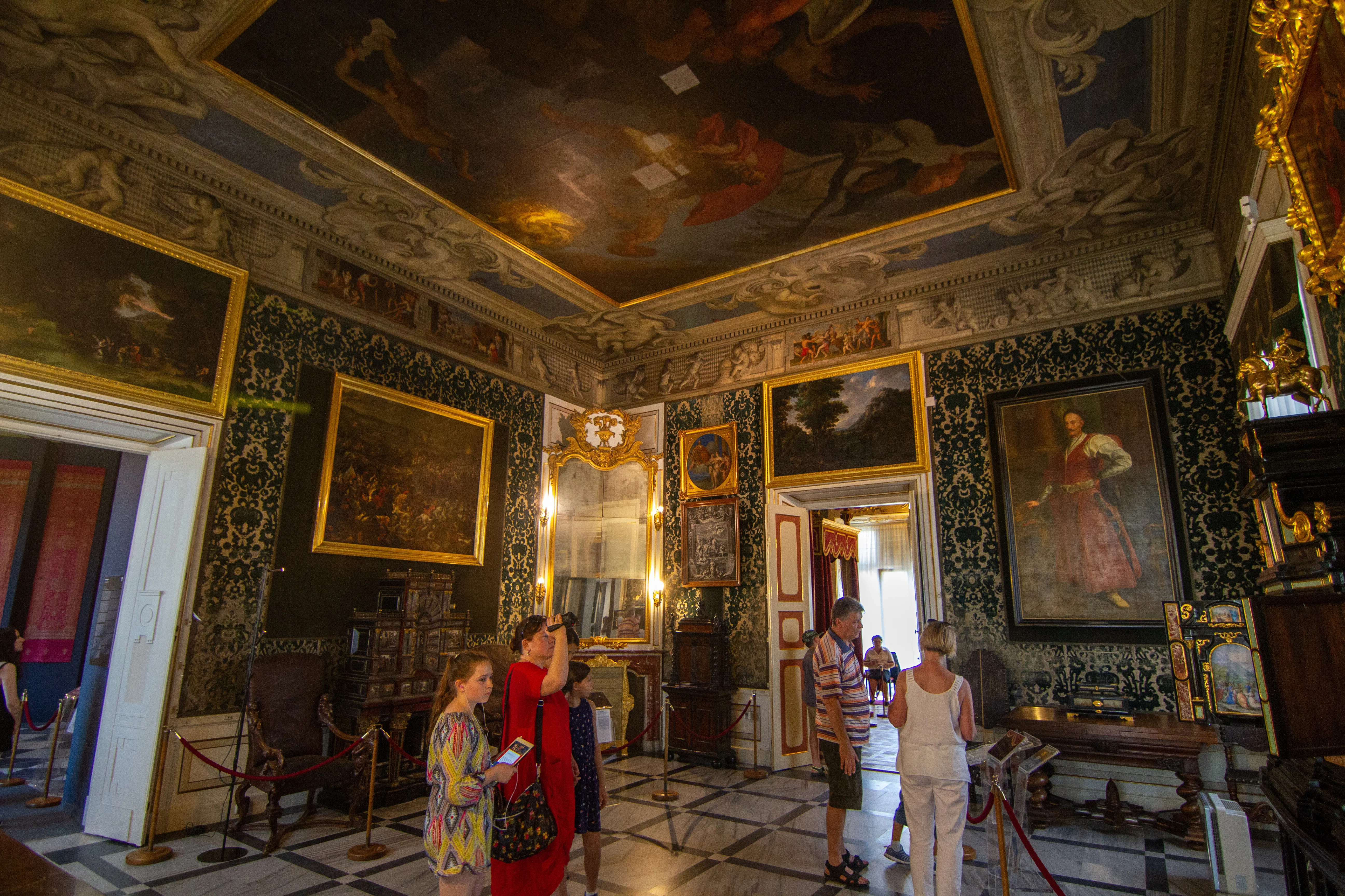 Ornada sala barroca con techo pintado, marcos dorados y visitantes