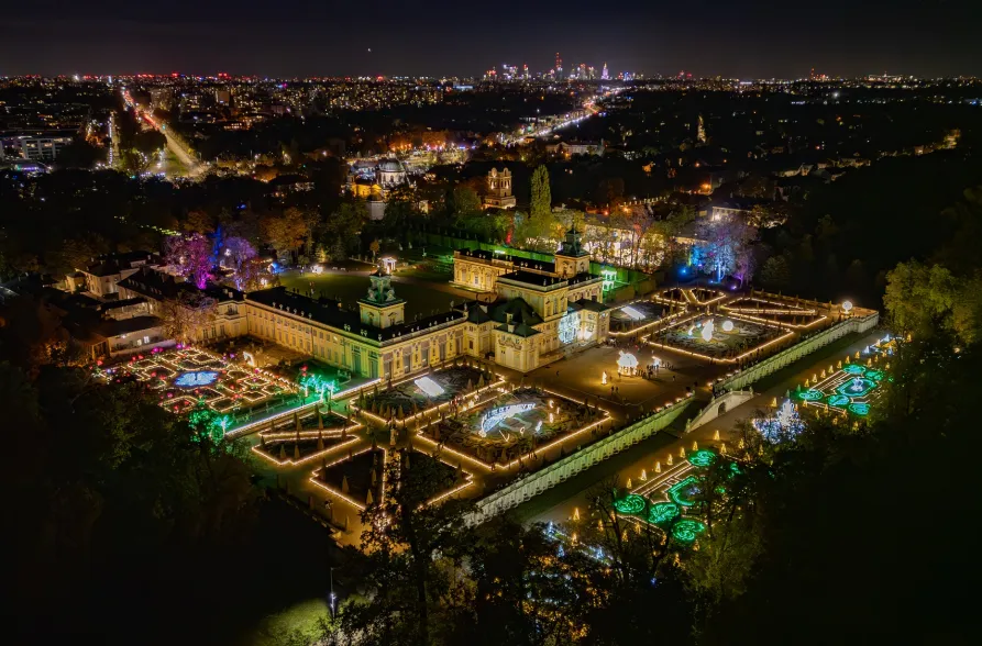 Vista aérea nocturna del Palacio de Wilanów iluminado con luces de colores, rodeado de jardines formales y el paisaje urbano