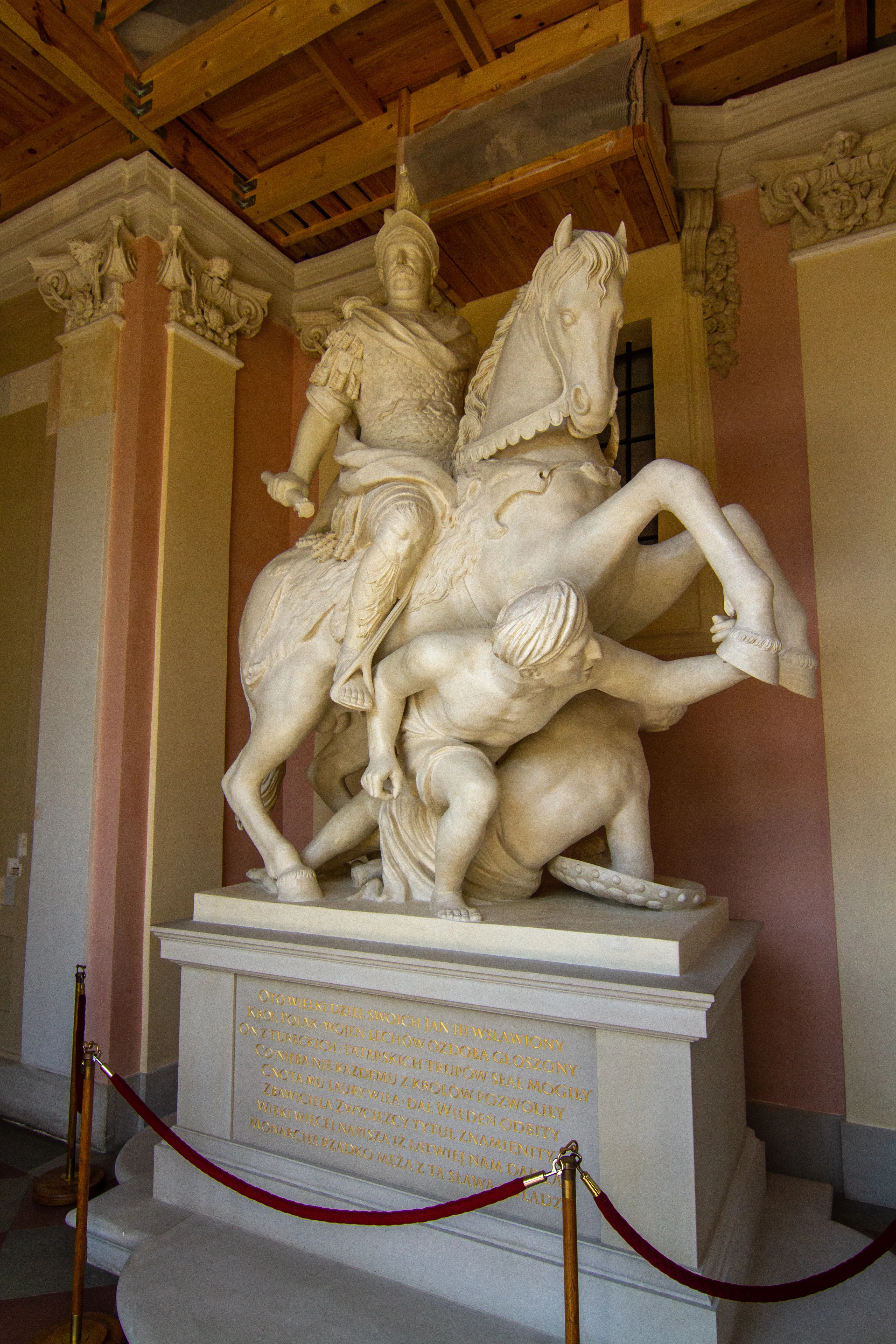 Marble sculpture of a mounted figure on a rearing horse above a fallen man, displayed in a palace hall with ornate columns and a wooden ceiling.