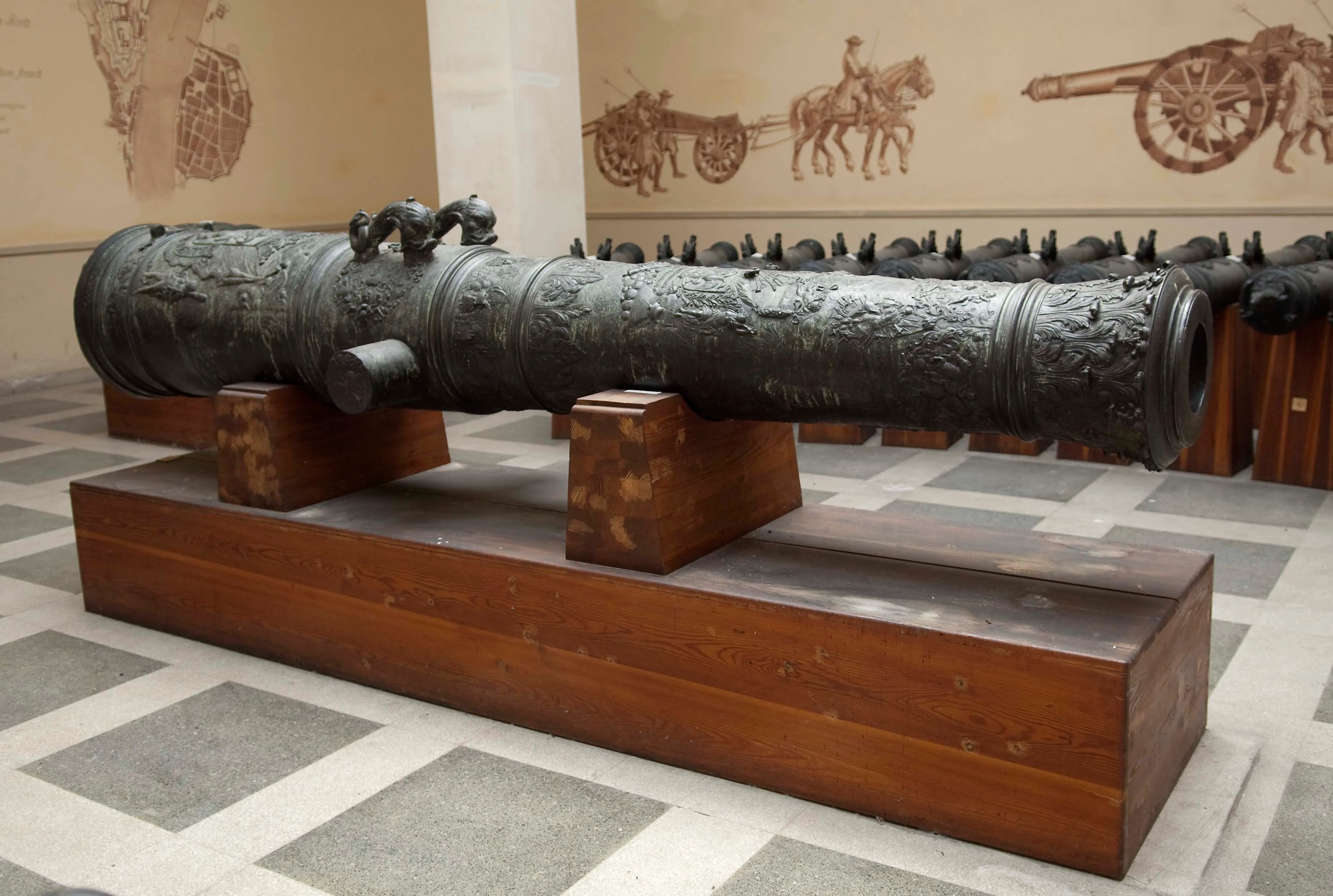 Een groot, sierlijk kanon op een houten voetstuk in een museumgalerij met historische muurschilderingen aan de muren