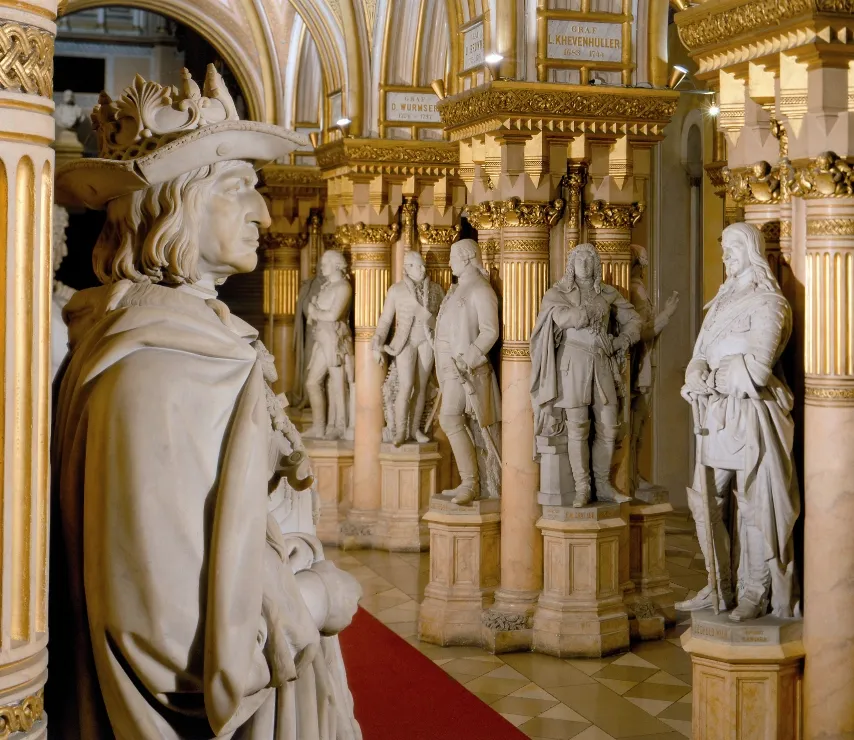 Marmeren beelden van historische figuren in een statige zaal met gouden zuilen en een rood tapijt