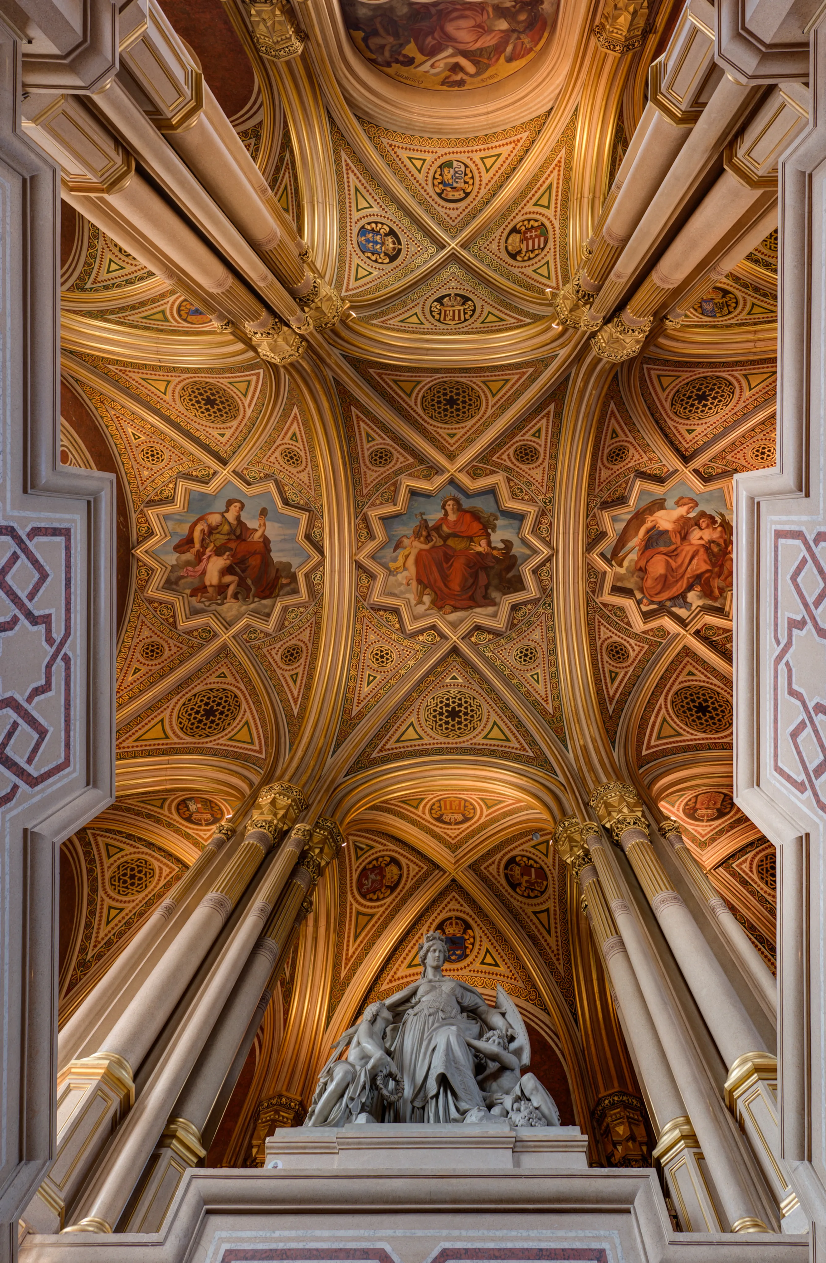 Gouden en crèmekleurige decoratieve patronen op een gewelfd plafond, fresco's en een centraal standbeeld in een grote hal