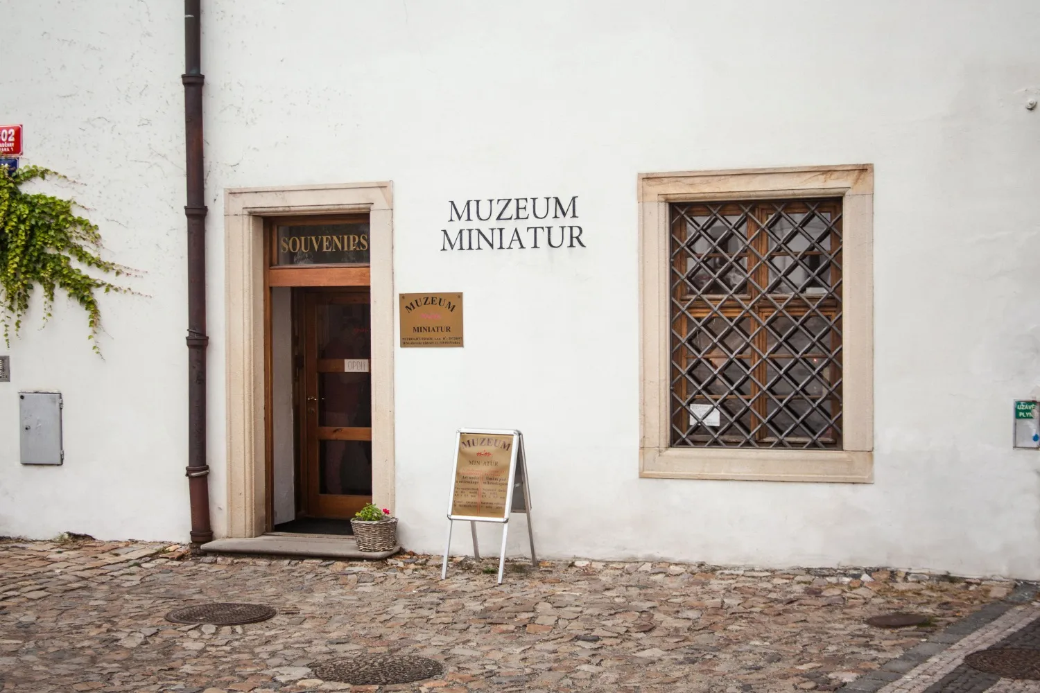 Fachada de edificio blanco con una puerta de madera, un cartel que dice 'MUZEUM MINIATUR' y una ventana con reja de metal.