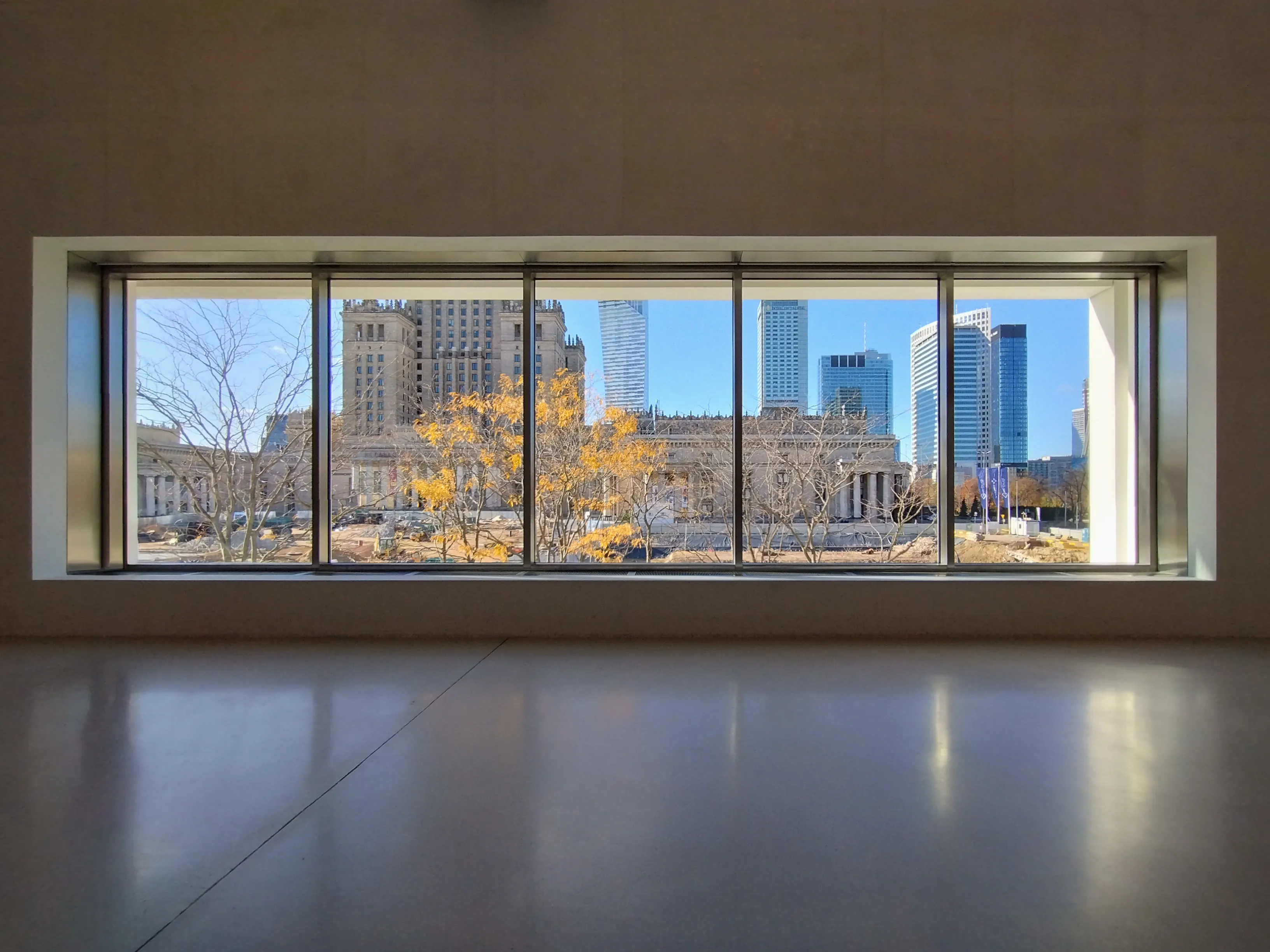 Large window in a modern interior overlooking a cityscape with buildings and trees with yellow leaves.