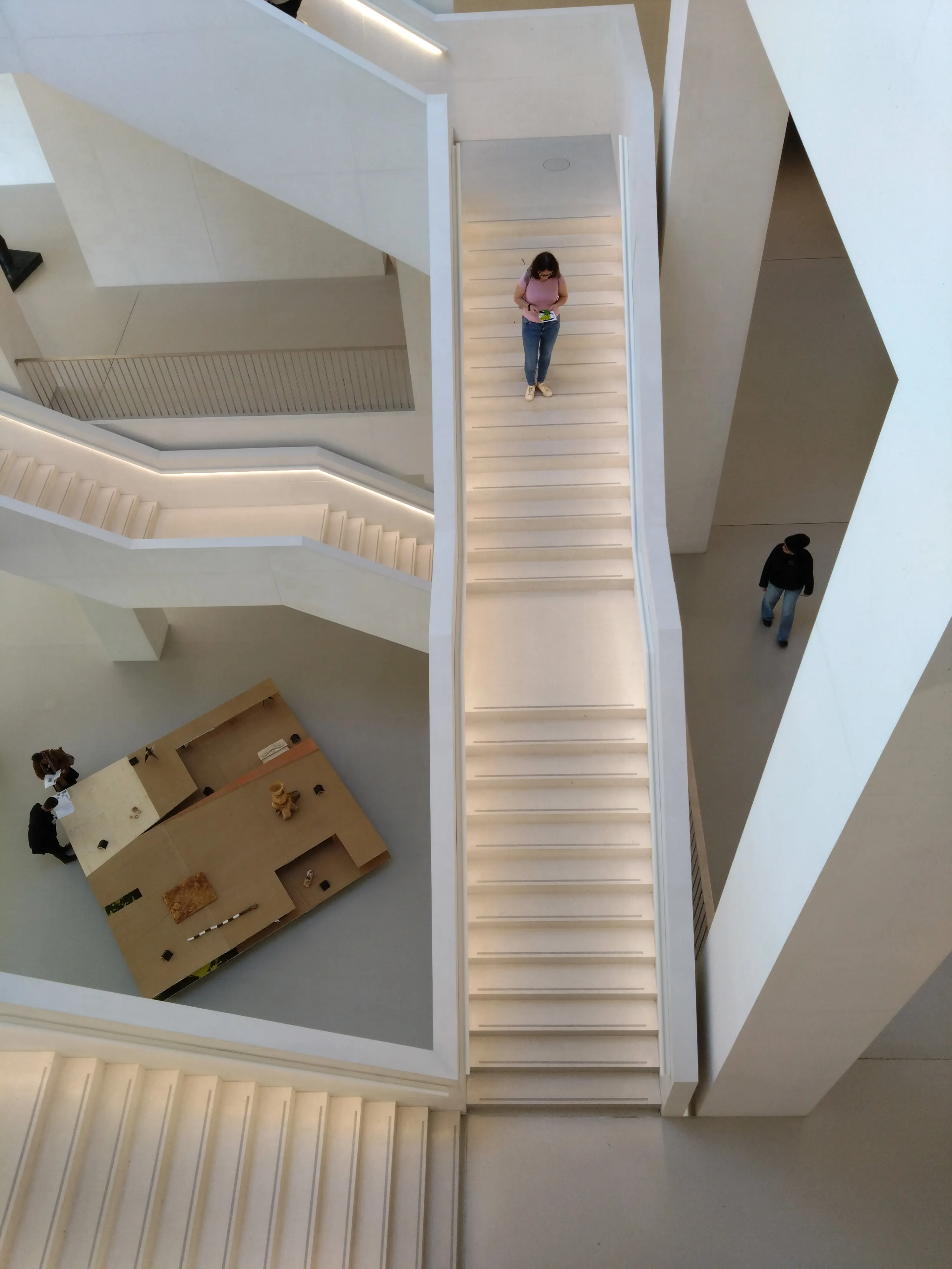 High-angle view of a modern white staircase in a museum interior with visitors walking on the steps and a cardboard installation on the floor below
