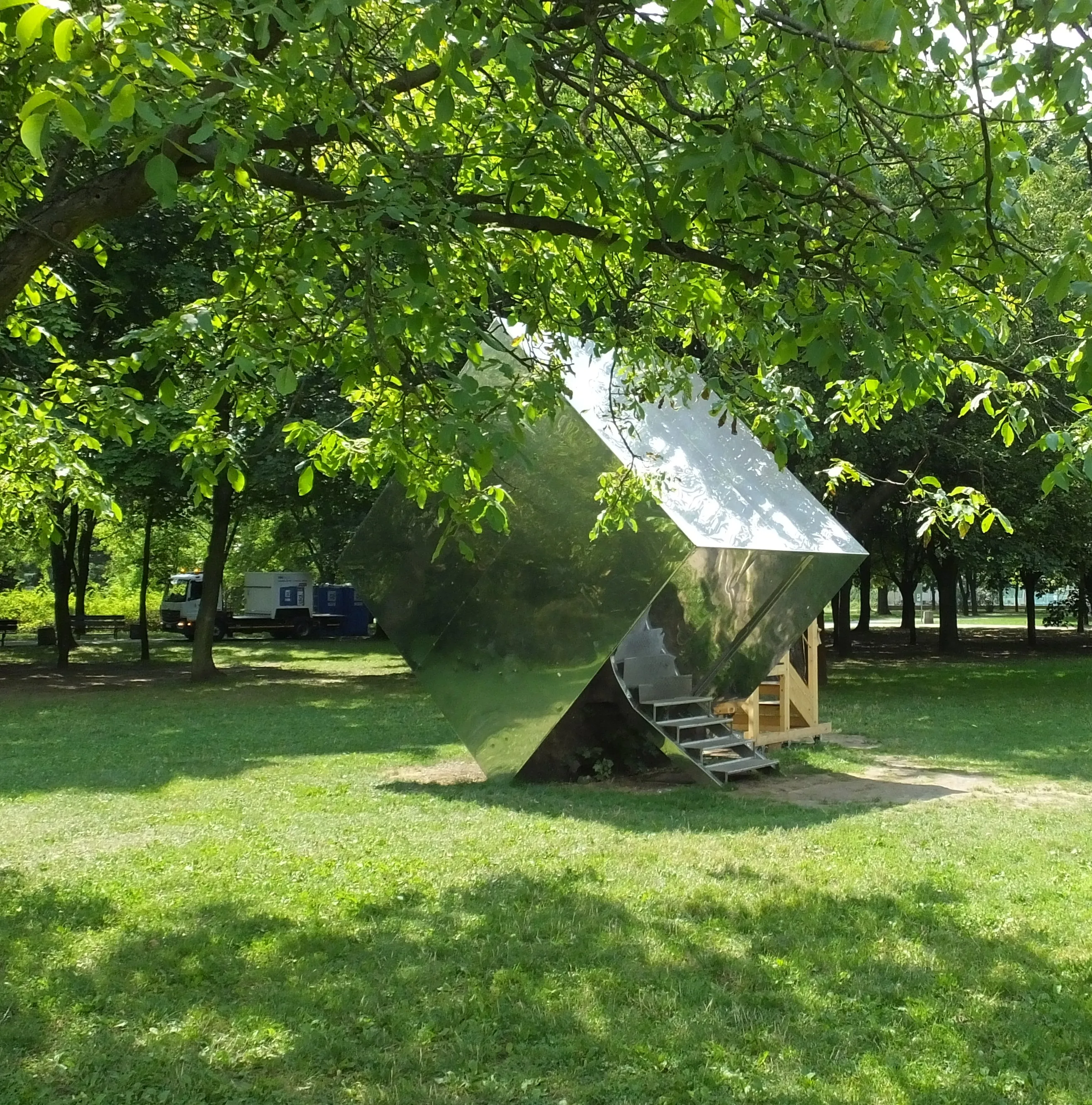 A modern metallic sculpture in a park with trees and grass