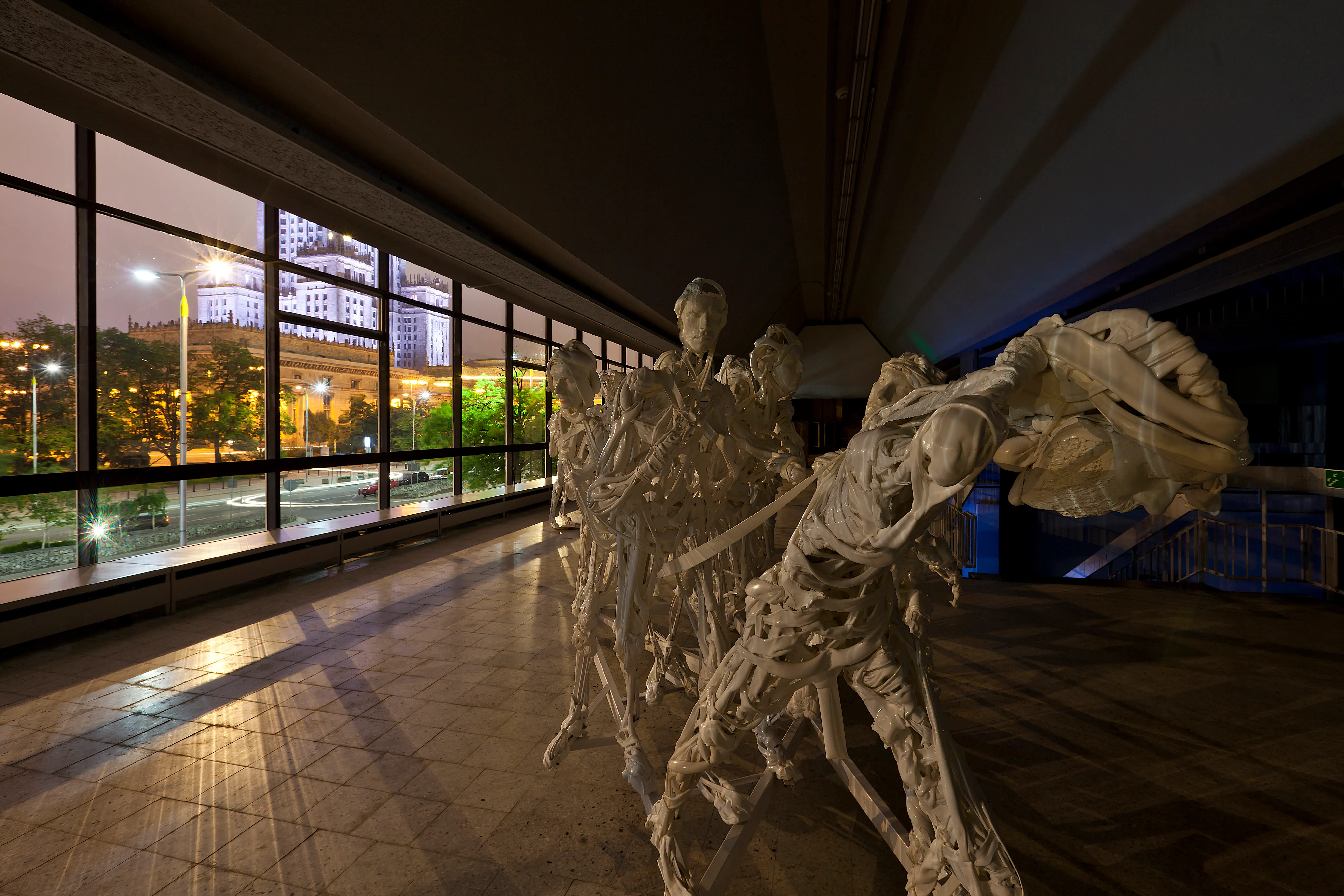 A large white sculpture of multiple figures in dynamic poses inside a gallery with a long window showing a city at night