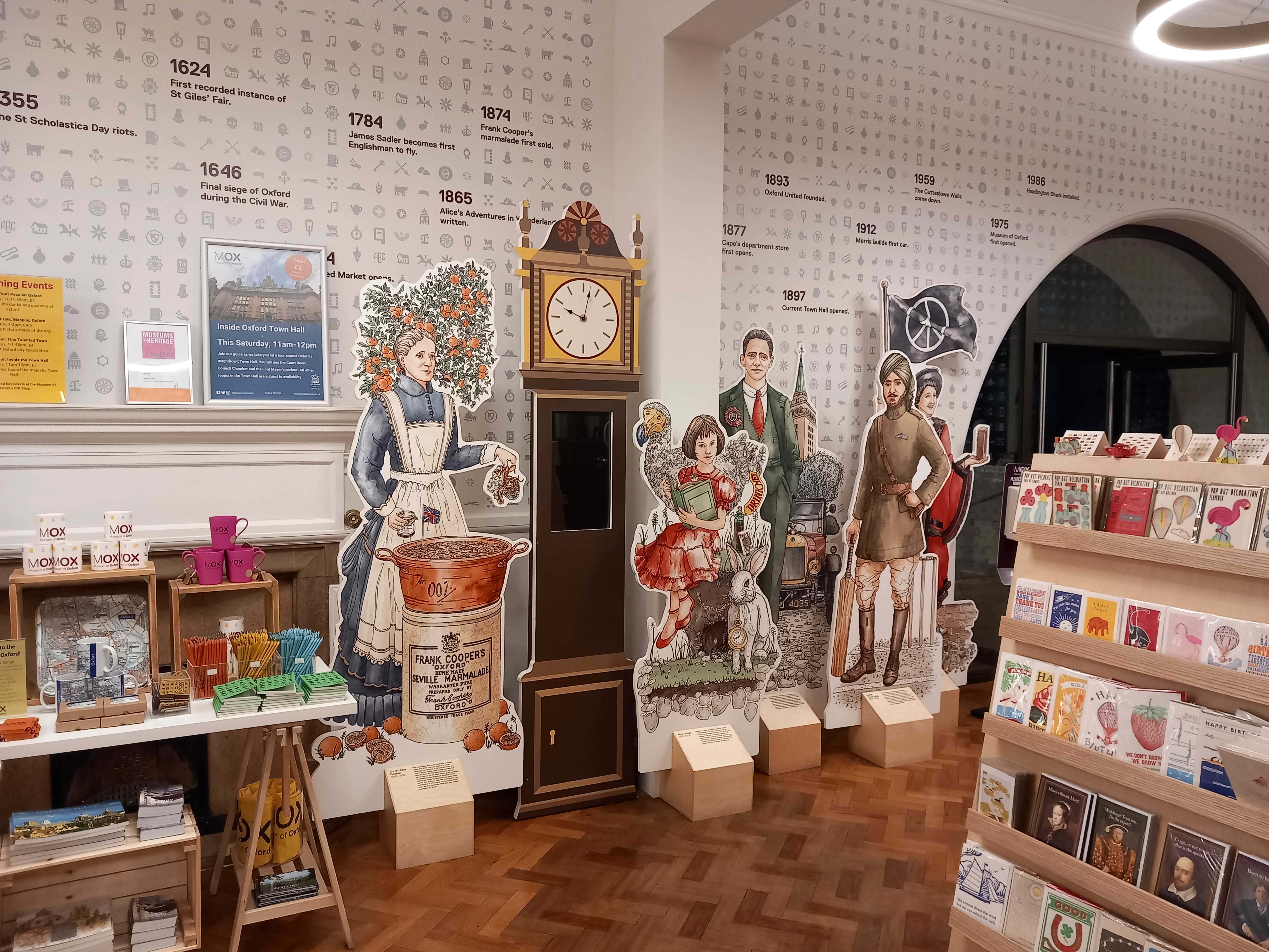 Interior de la tienda de regalos del Museum of Oxford con siluetas de cartón de figuras históricas, un reloj de pie, estantes de mercancía y una pared con una cronología y fechas.