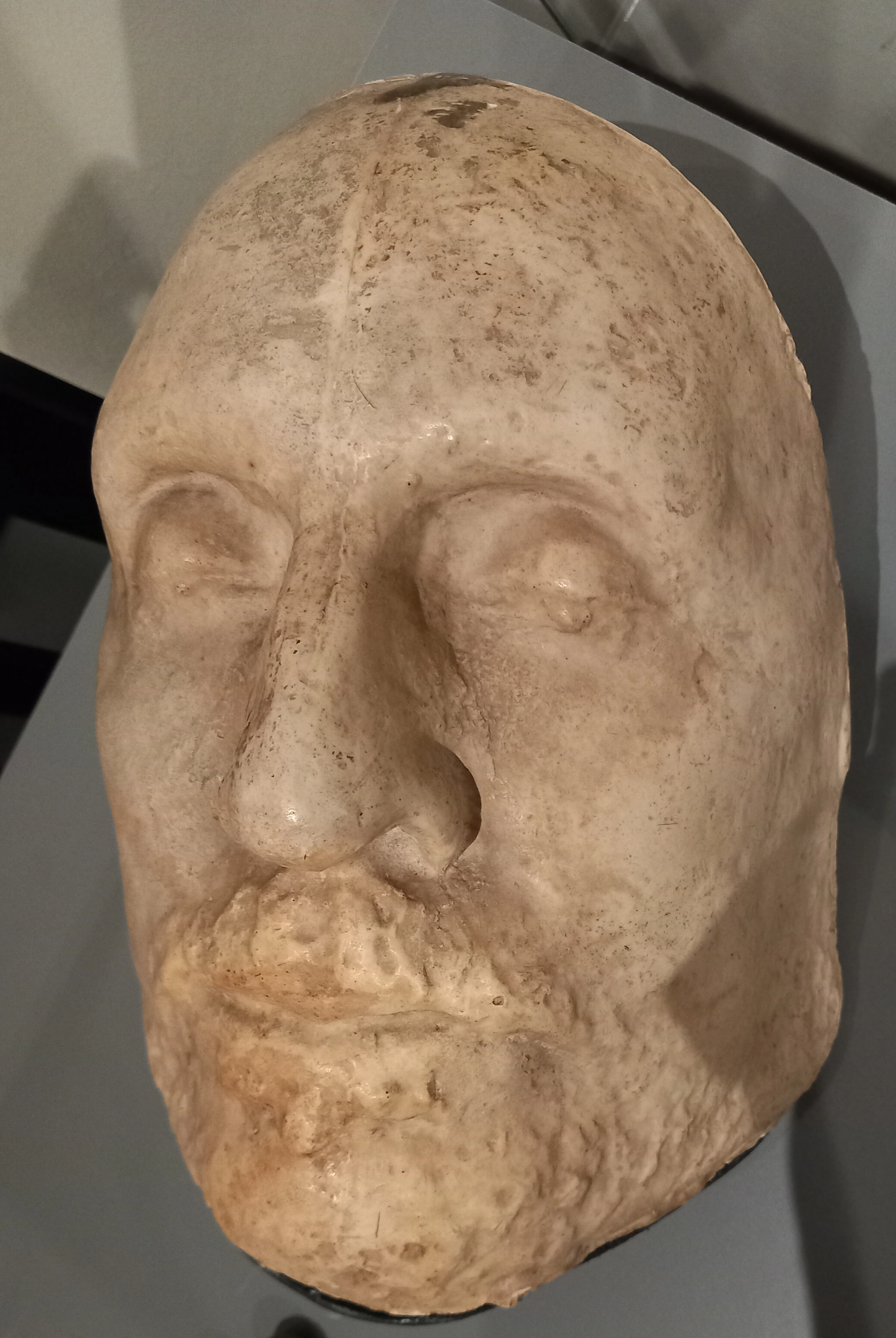 Máscara mortuoria de yeso de un hombre barbudo con los ojos cerrados y superficie texturizada, expuesta sobre un pedestal gris de museo.